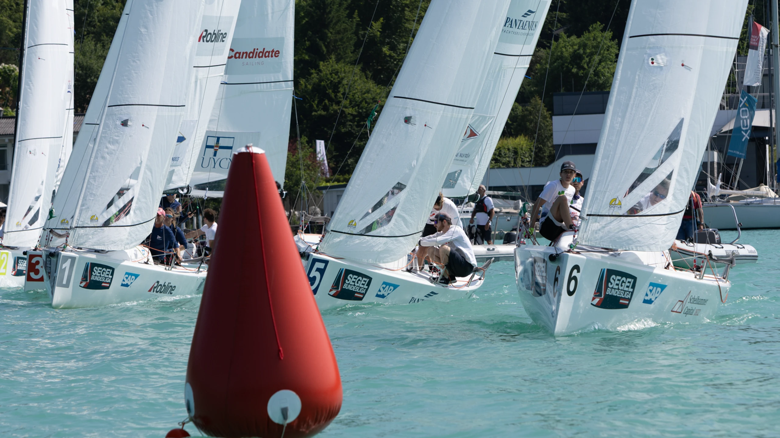 Segelregatta auf dem See mit mehreren Jollen unter weißen Spinnakern, eine rote Regattatonne im Vordergrund.