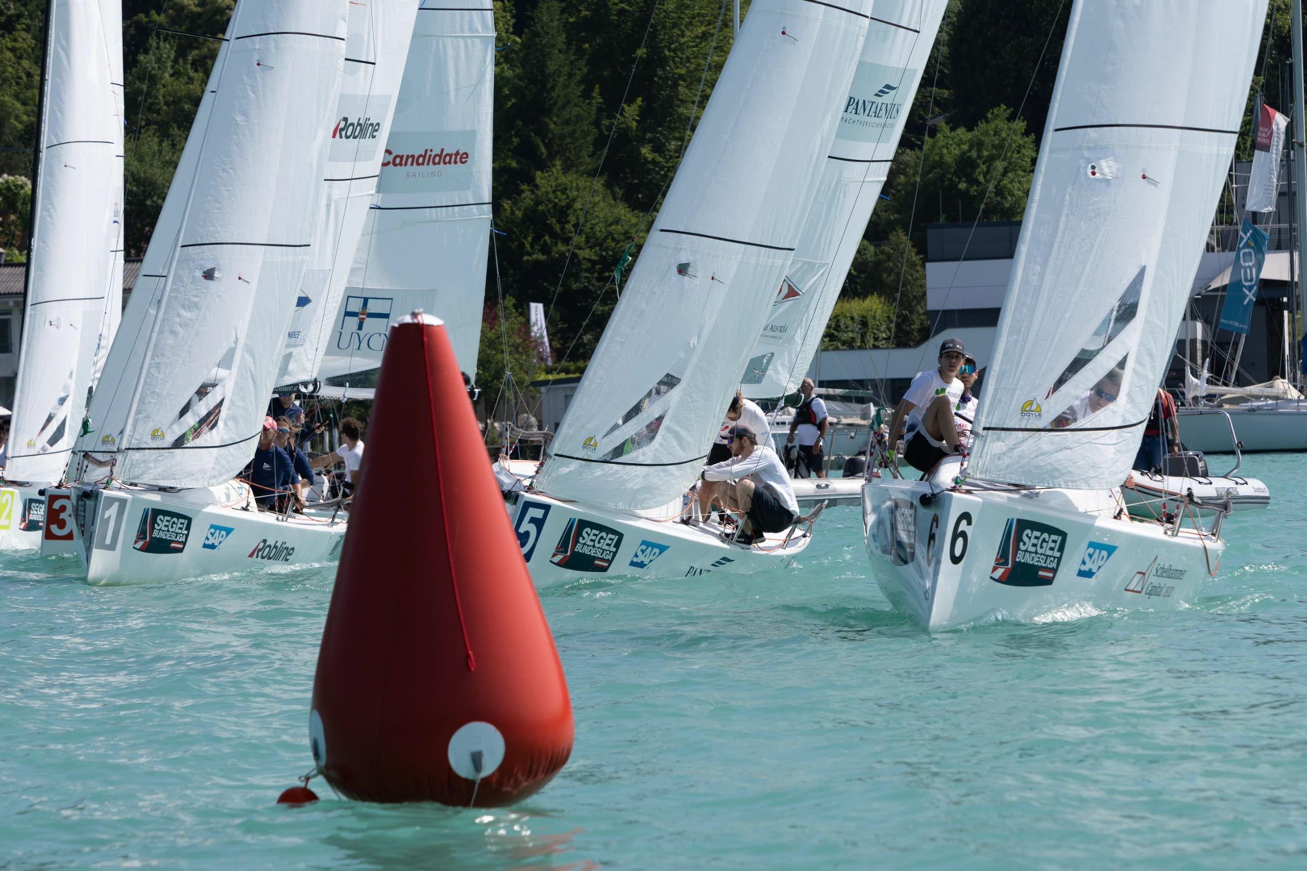Segelregatta auf dem See mit mehreren Jollen unter weißen Spinnakern, eine rote Regattatonne im Vordergrund.