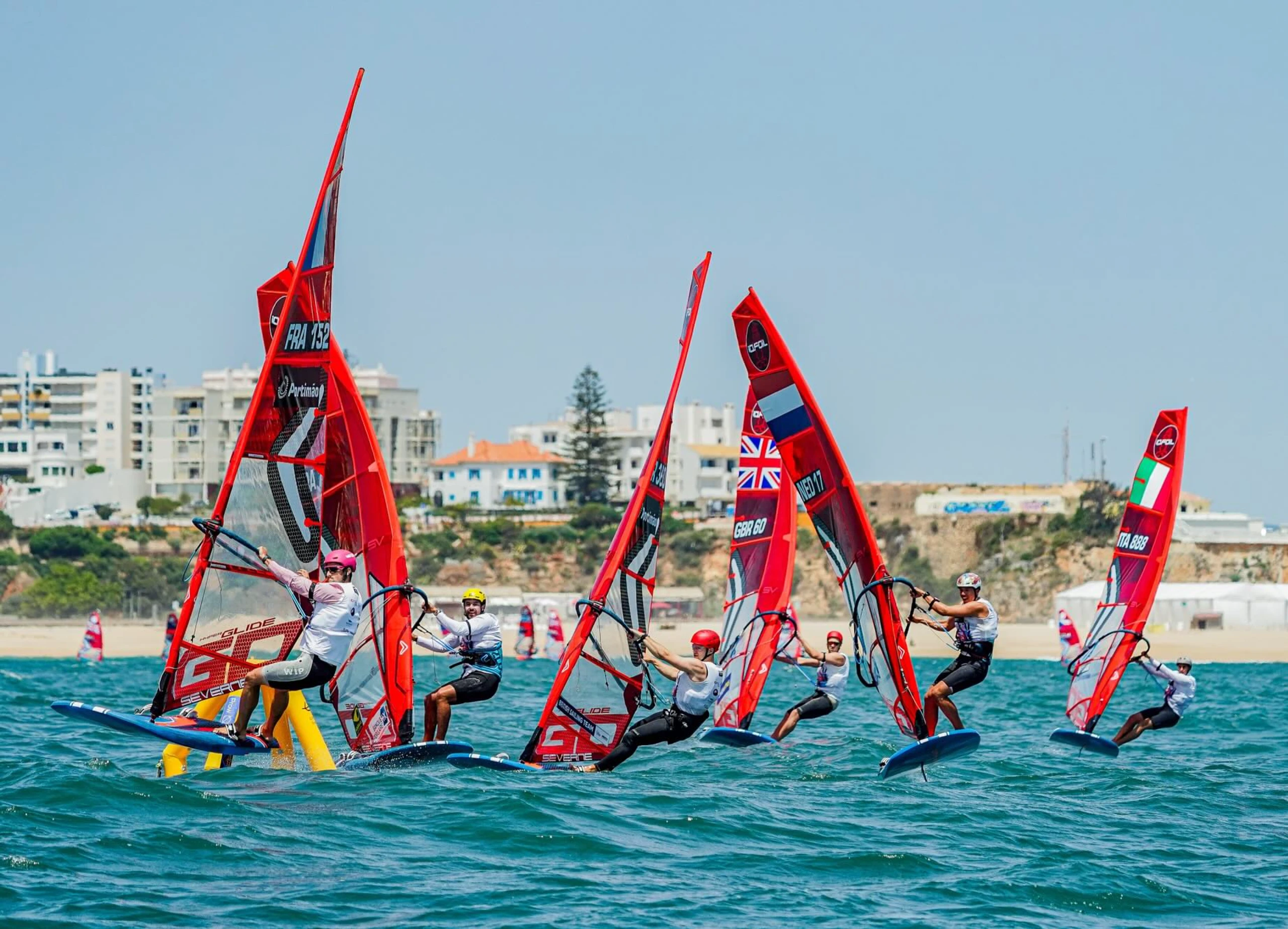 Sechs Windsurfer mit roten Segeln vor der portugiesischen Küste fahren um eine gelbe Markierung im Wasser.
