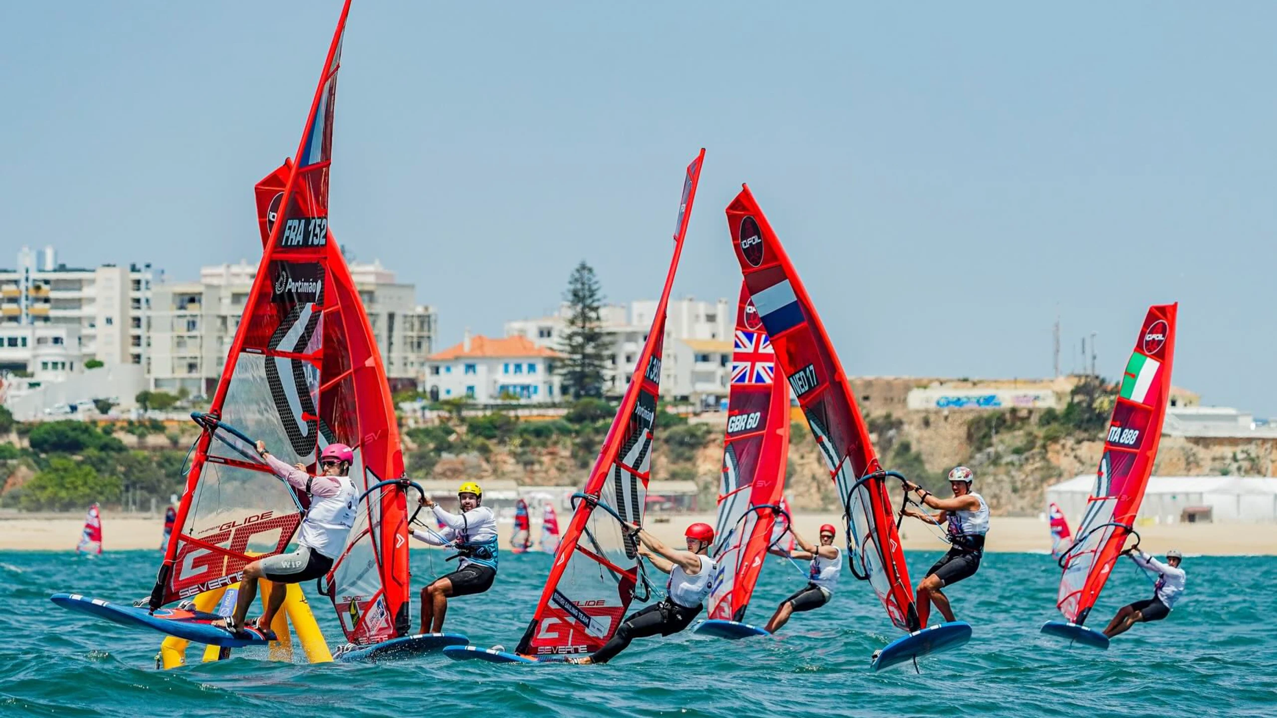 Sechs Windsurfer mit roten Segeln vor der portugiesischen Küste fahren um eine gelbe Markierung im Wasser.