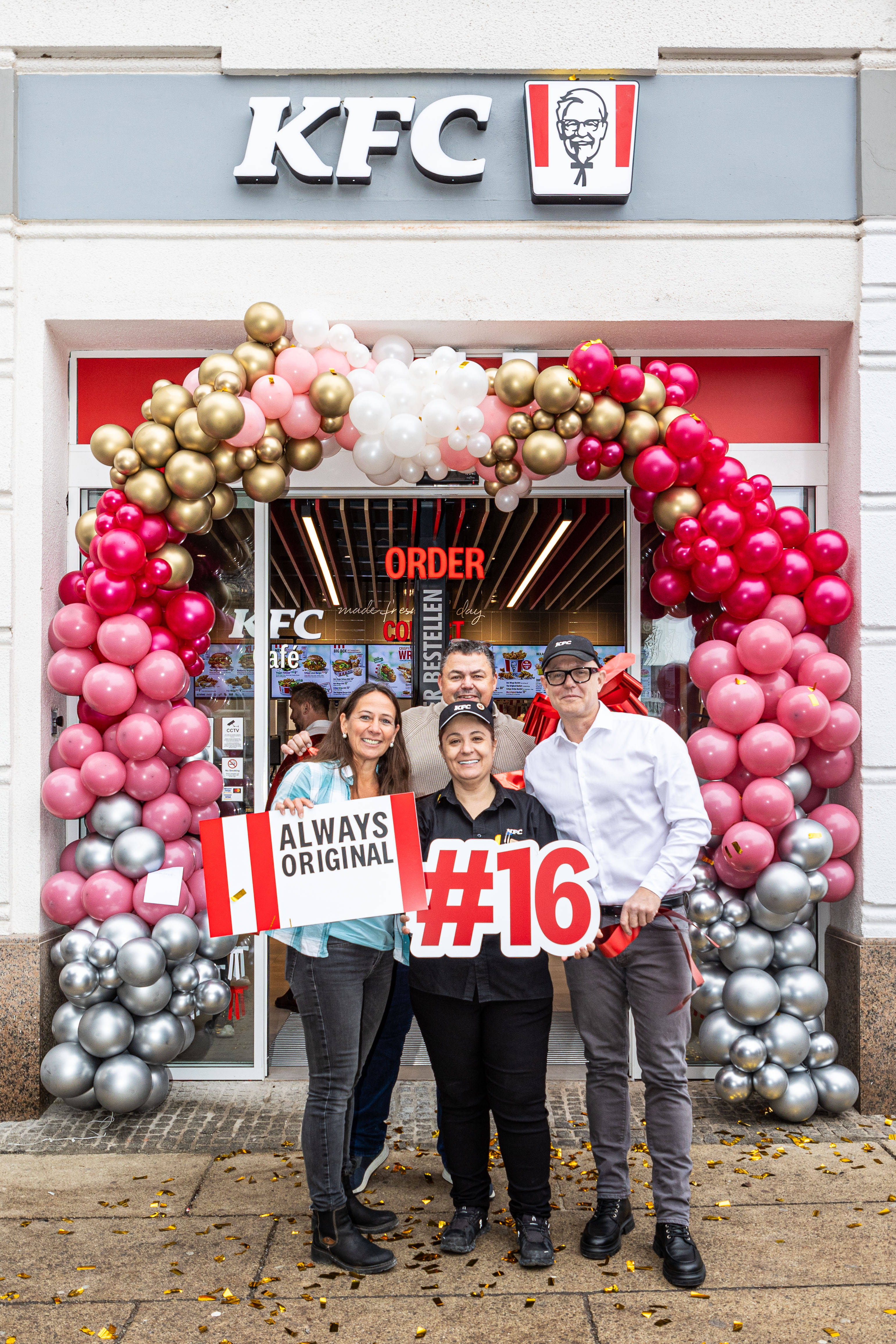 Team hält Schild '#16' vor der KFC-Filiale St. Pölten, Eingang mit Ballongirlande geschmückt