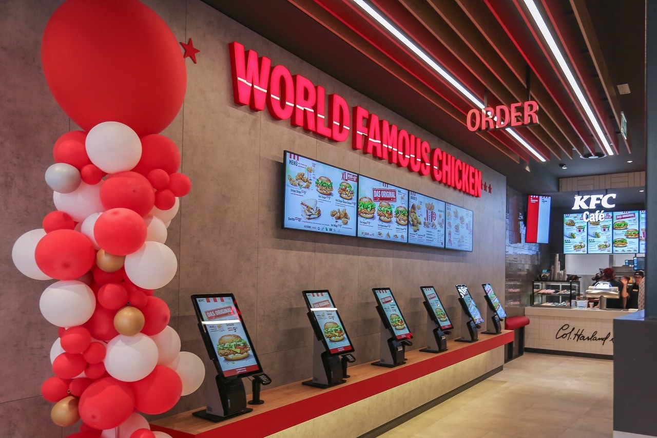 Bestellkiosks in einer KFC-Filiale mit Leuchtschriftzug 'World Famous Chicken' und Ballondeko