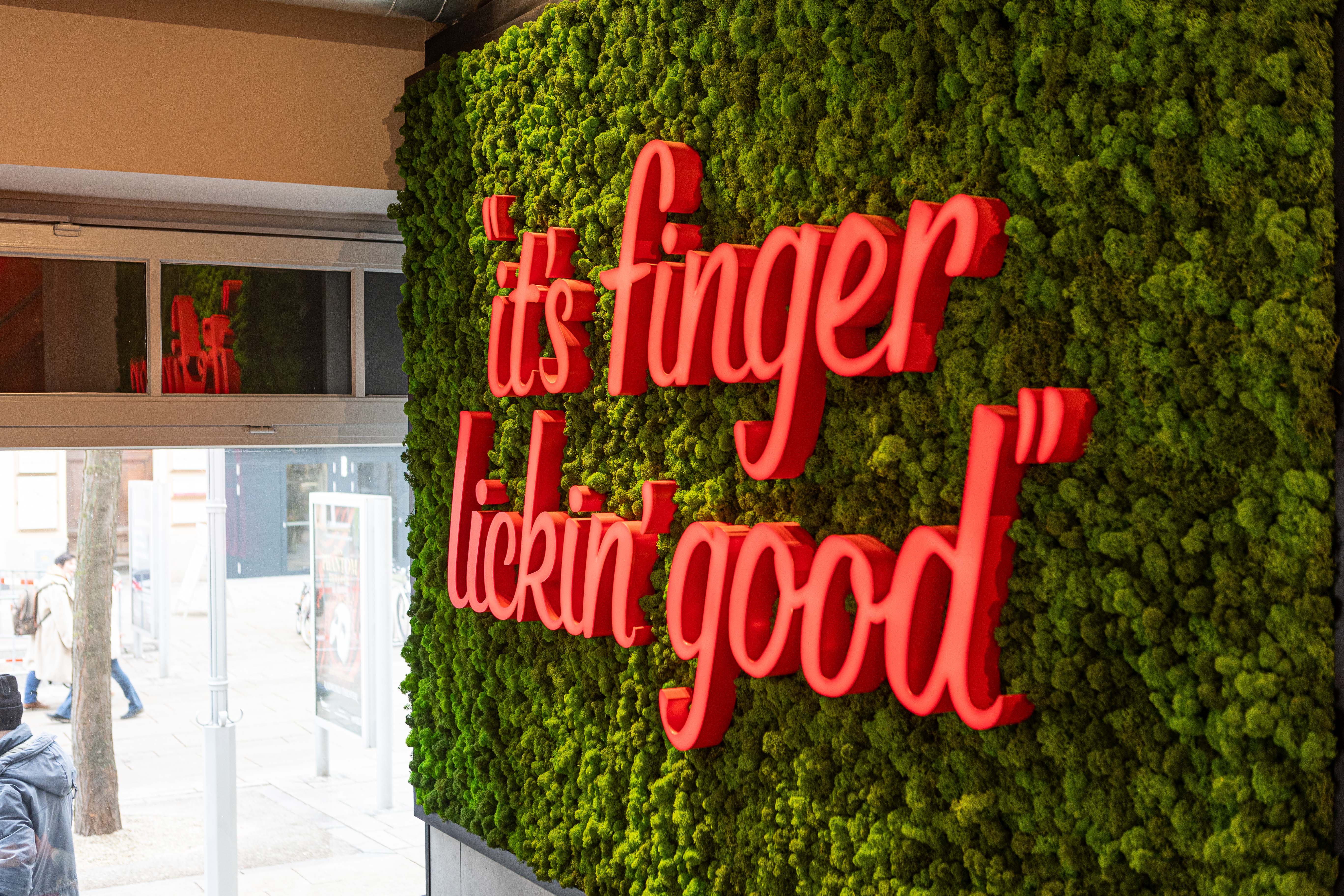 Moosbegrünte Wand mit rotem Leuchtschriftzug 'It's Finger Lickin' Good' in der Filiale St. Pölten