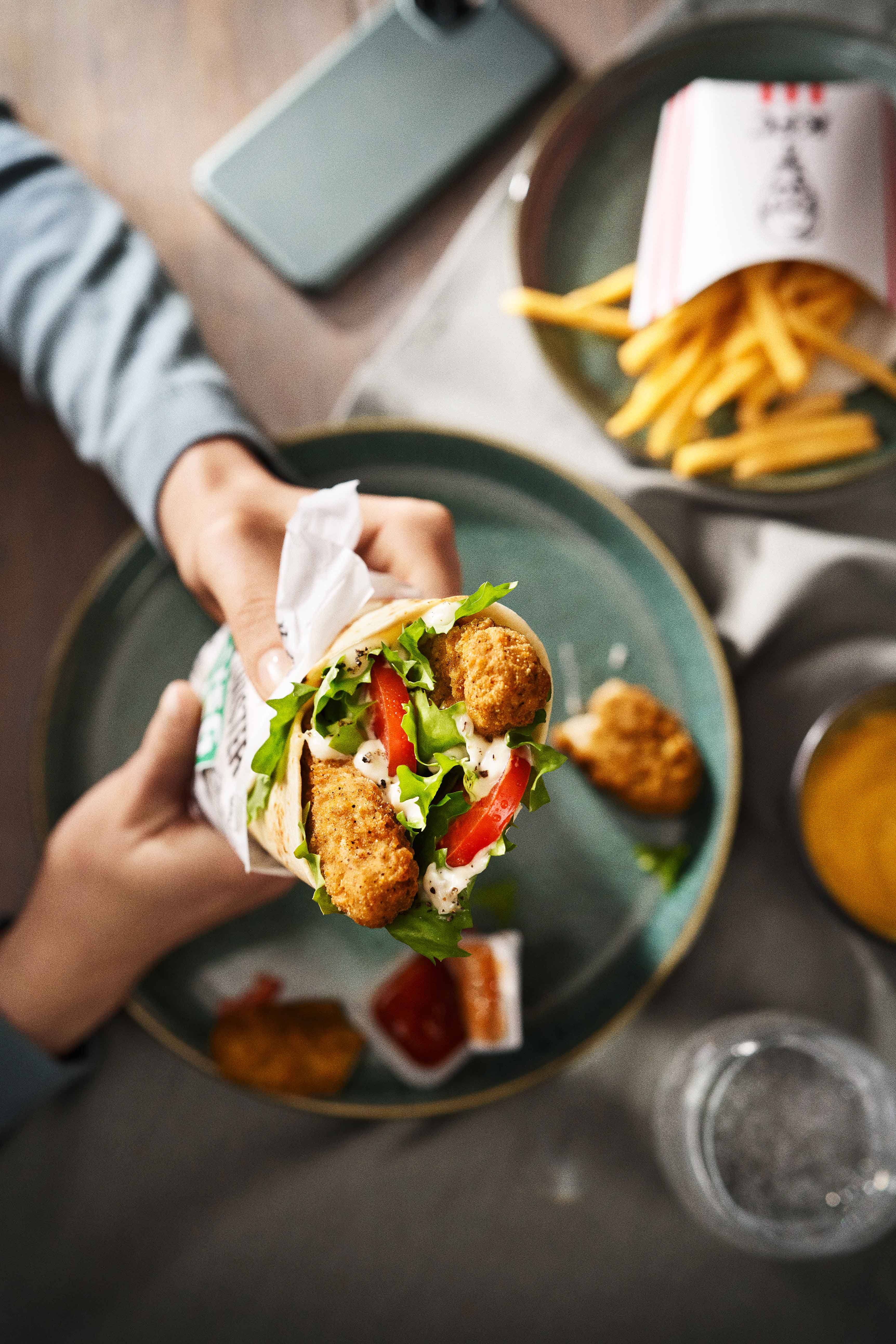 Hände halten KFC Veggie Twister Wrap, daneben Pommes und Dips