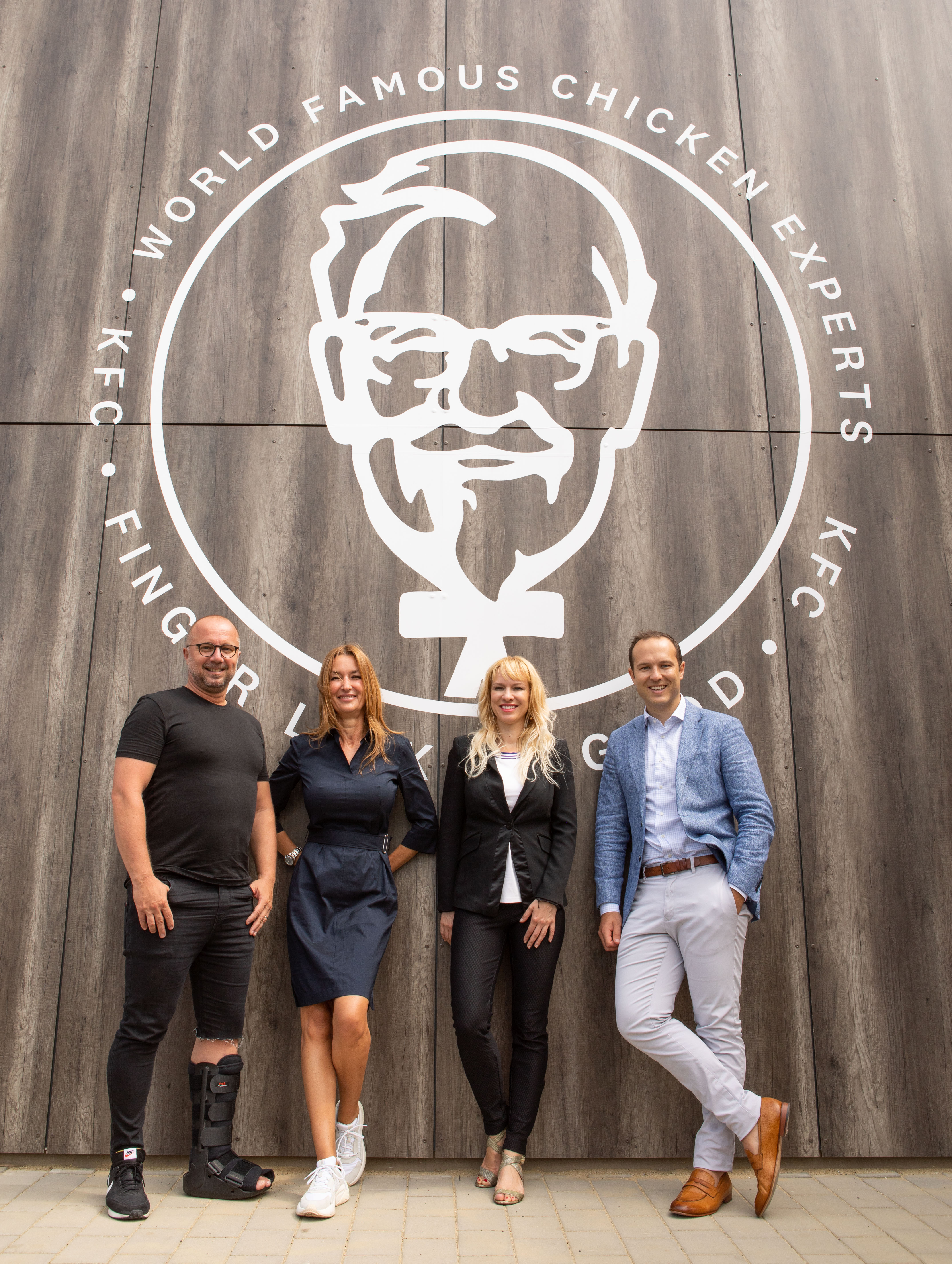 Vier Personen vor einer Wand mit Colonel-Sanders-Illustration und Schriftzug 'World Famous Chicken Experts'