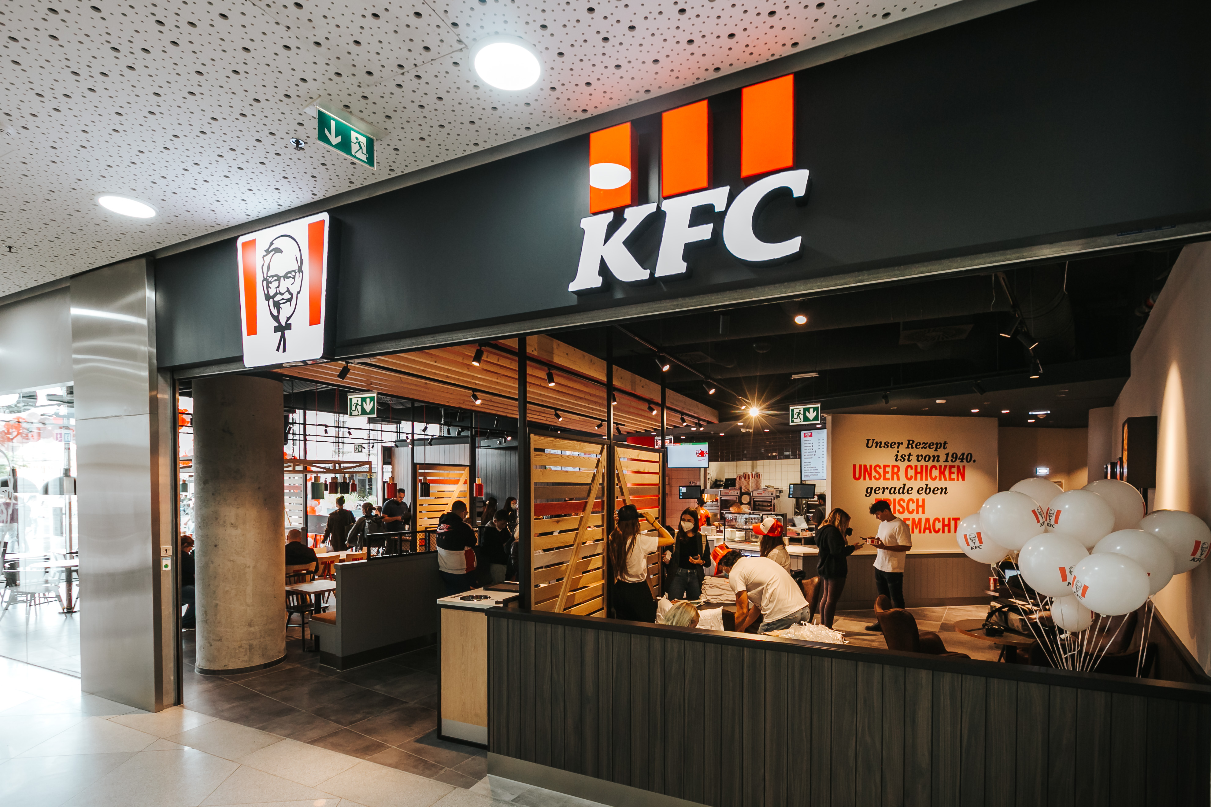 Eingang einer KFC-Filiale mit Leuchtschrift, Werbeplakat und Luftballons zur Neueröffnung