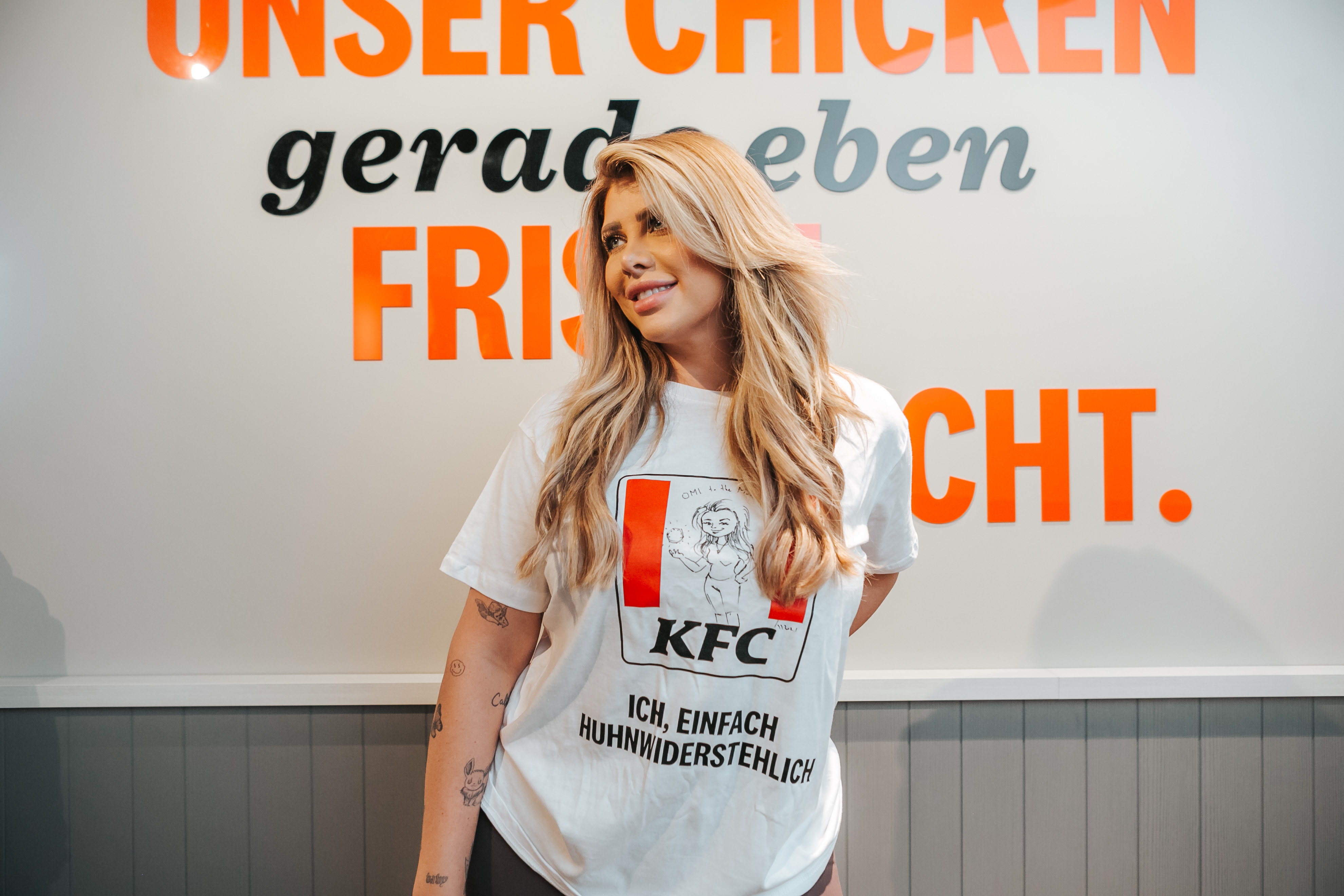 Frau im weißen KFC-T-Shirt vor Wandschriftzug 'Unser Chicken gerade eben frisch gemacht'