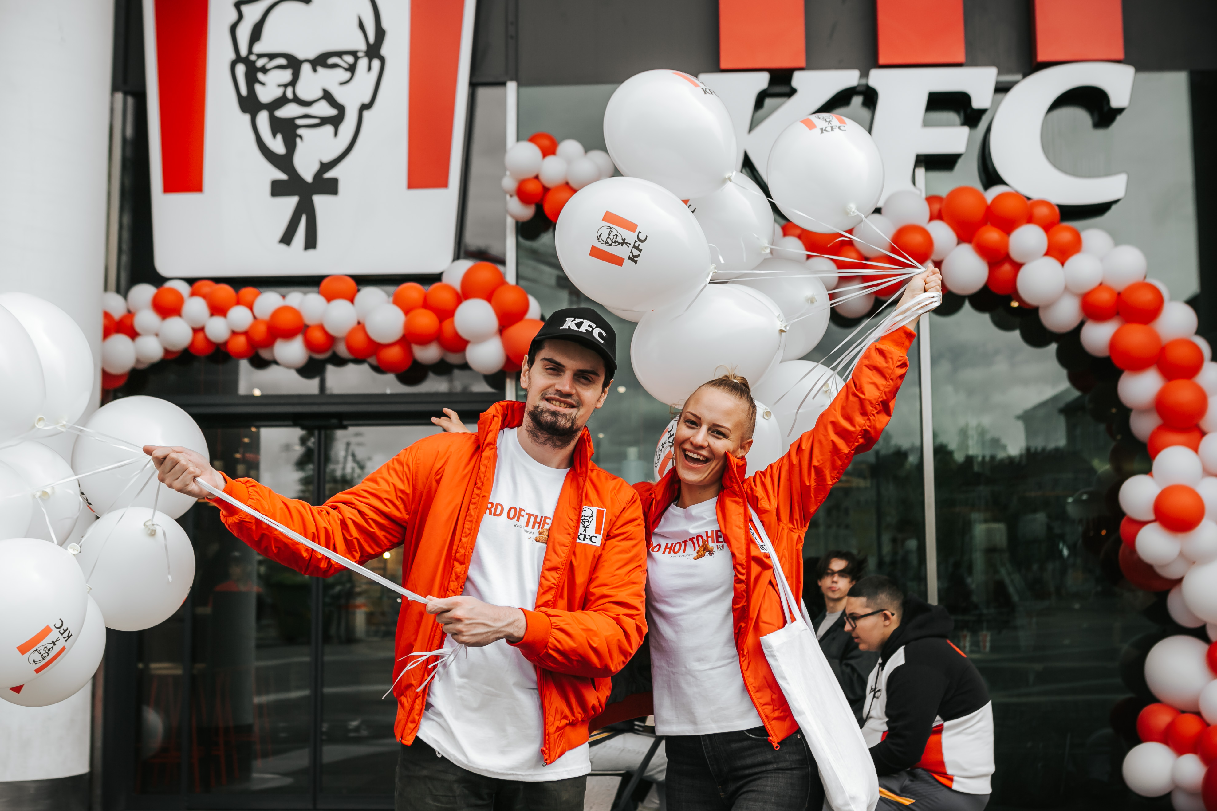 Zwei Personen in orangen KFC-Jacken mit Luftballons vor einer KFC-Filiale bei der Eröffnung