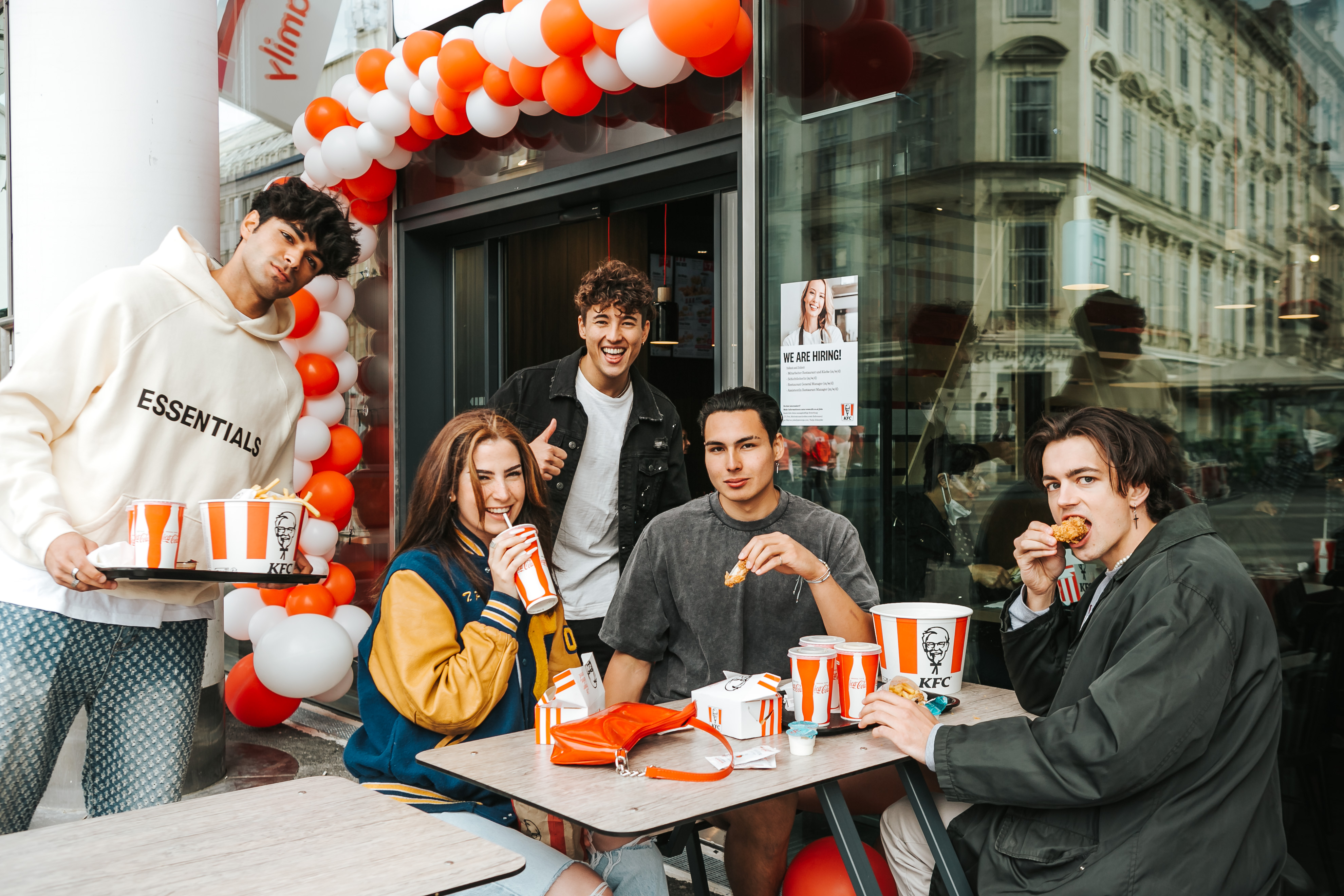 Gruppe junger Menschen isst KFC-Gerichte an einem Tisch vor der Filiale mit Ballongirlande