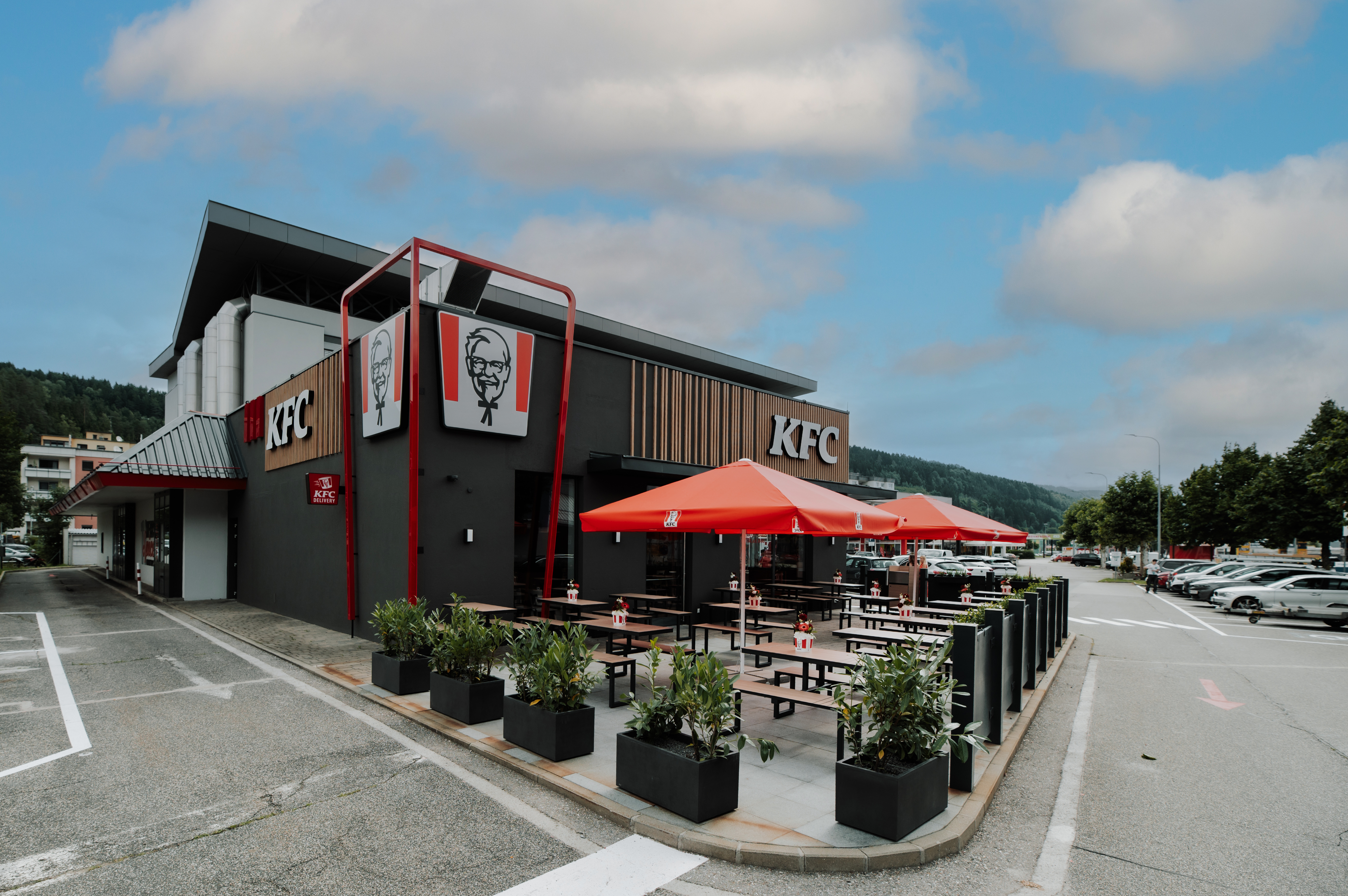 Außenterrasse einer KFC-Filiale mit roten Sonnenschirmen und Bergpanorama im Hintergrund