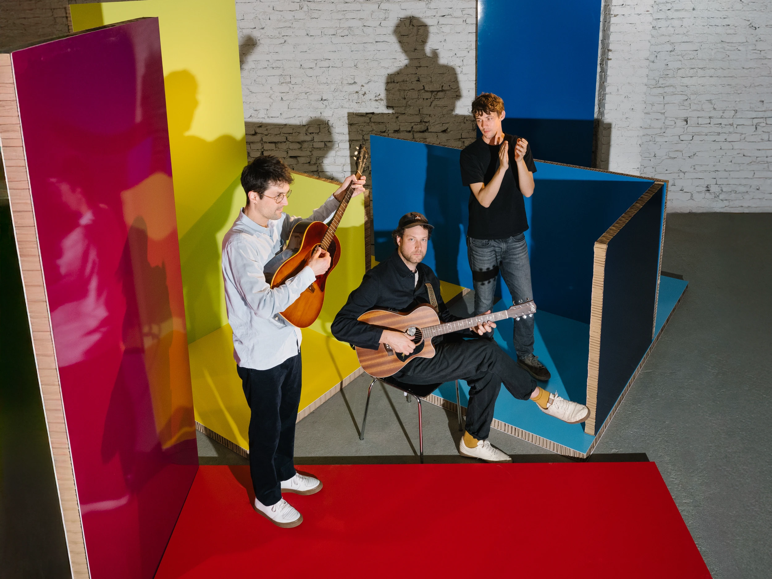 Drei Musiker mit Akustikgitarren vor einer bunten Kulisse aus roten, gelben und blauen Paneelen im Studio.