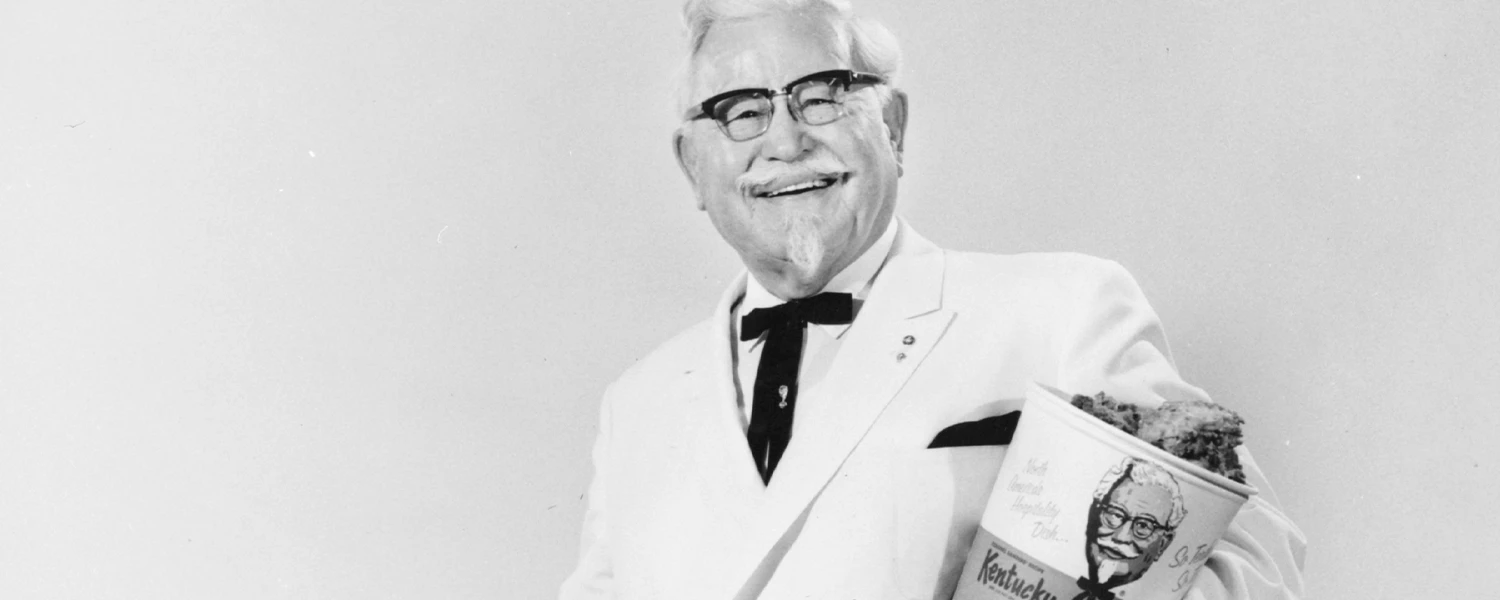 Colonel Harland Sanders im weissen Anzug mit schwarzer Fliege, lächelnd mit einem Kentucky-Fried-Chicken-Bucket im Arm