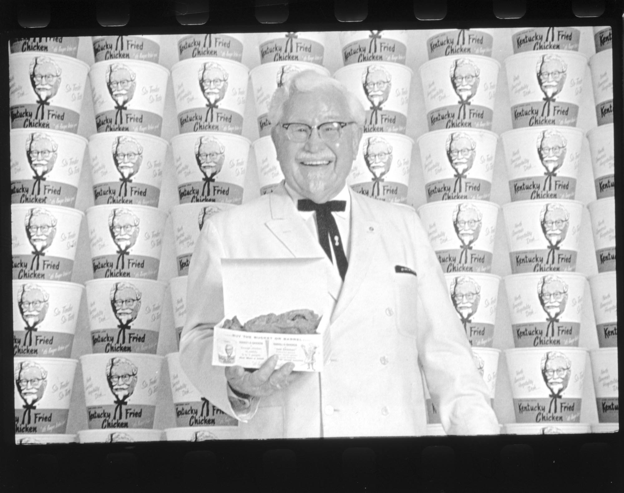 Plukovník Harland Sanders pred stenou z naskladaných vedierok Kentucky Fried Chicken
