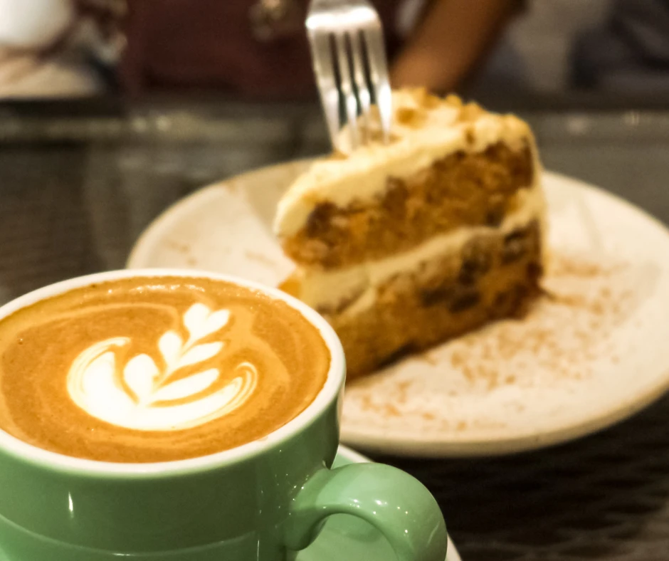 Eine Tasse Cappuccino mit Latte Art und ein Stück Kuchen mit Gabel auf einem Teller im Hintergrund.