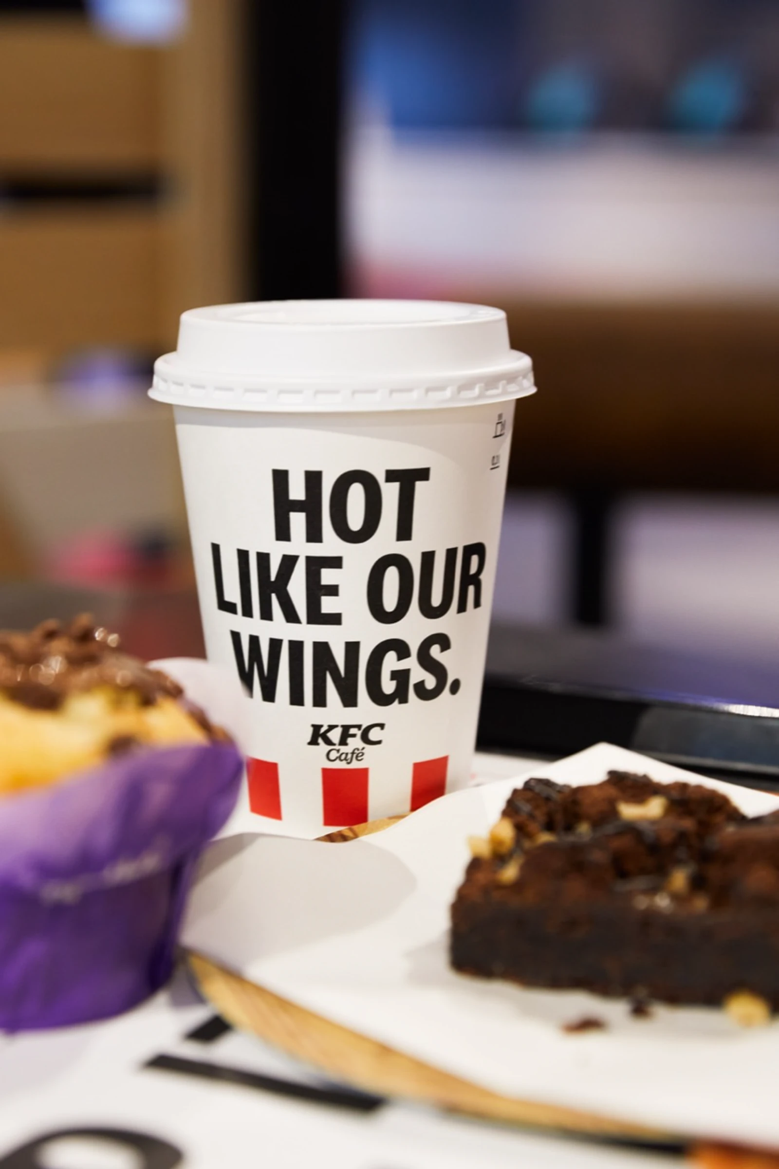 Pohár KFC Café s nápisom Hot like our wings, vedľa muffin a brownie