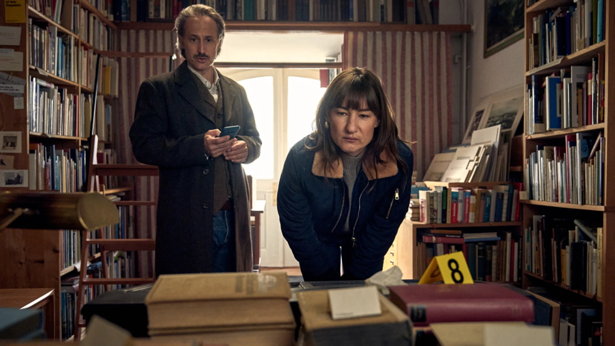 Michael Ostrowski und Marie Leuenberger inspizieren einen Tatort umringt von Bücherregalen