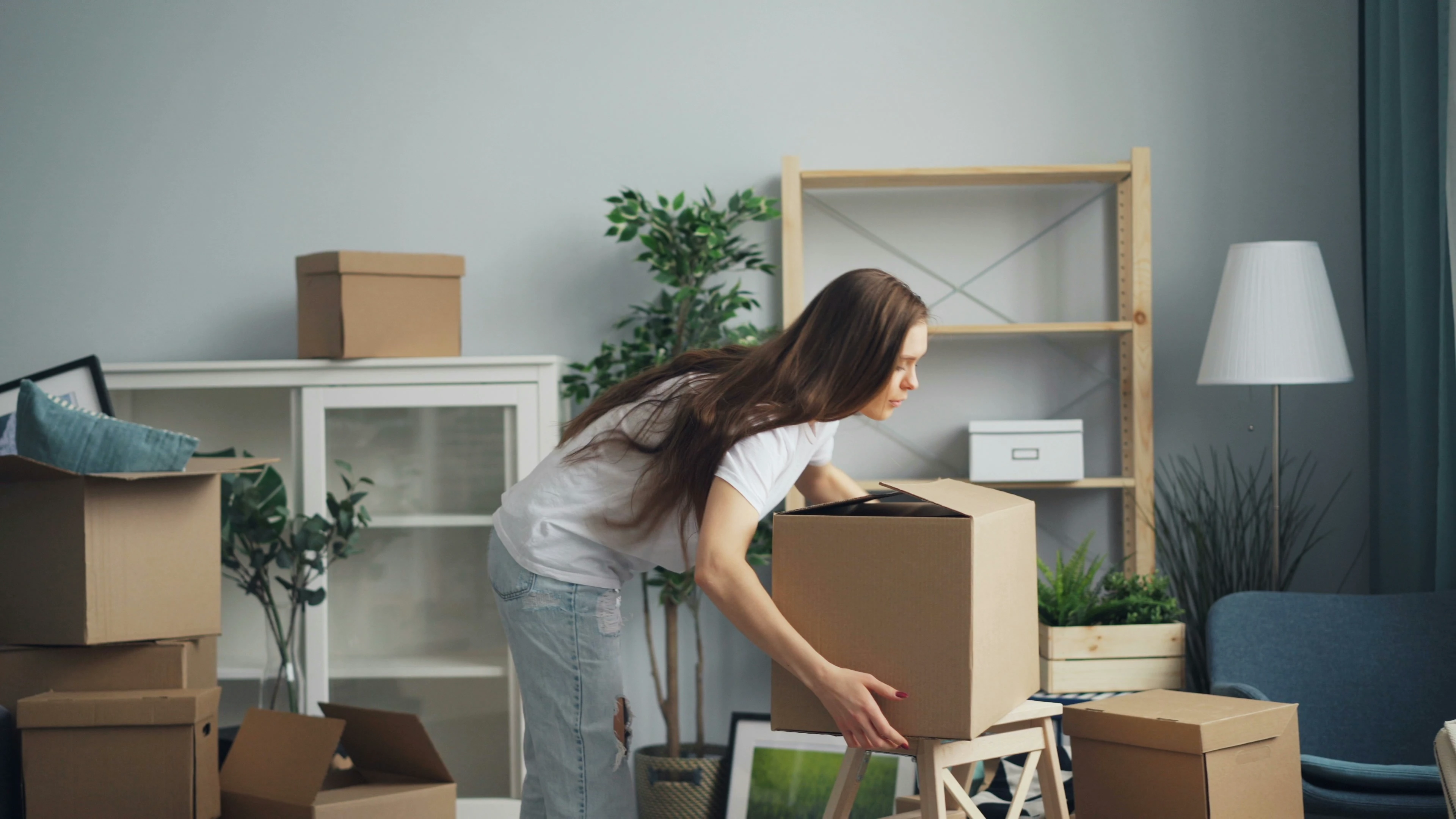 Eine junge Frau beim Ein- oder Auspacken von Umzugskartons in einem neuen Wohnzimmer mit Regalen und Pflanzen.