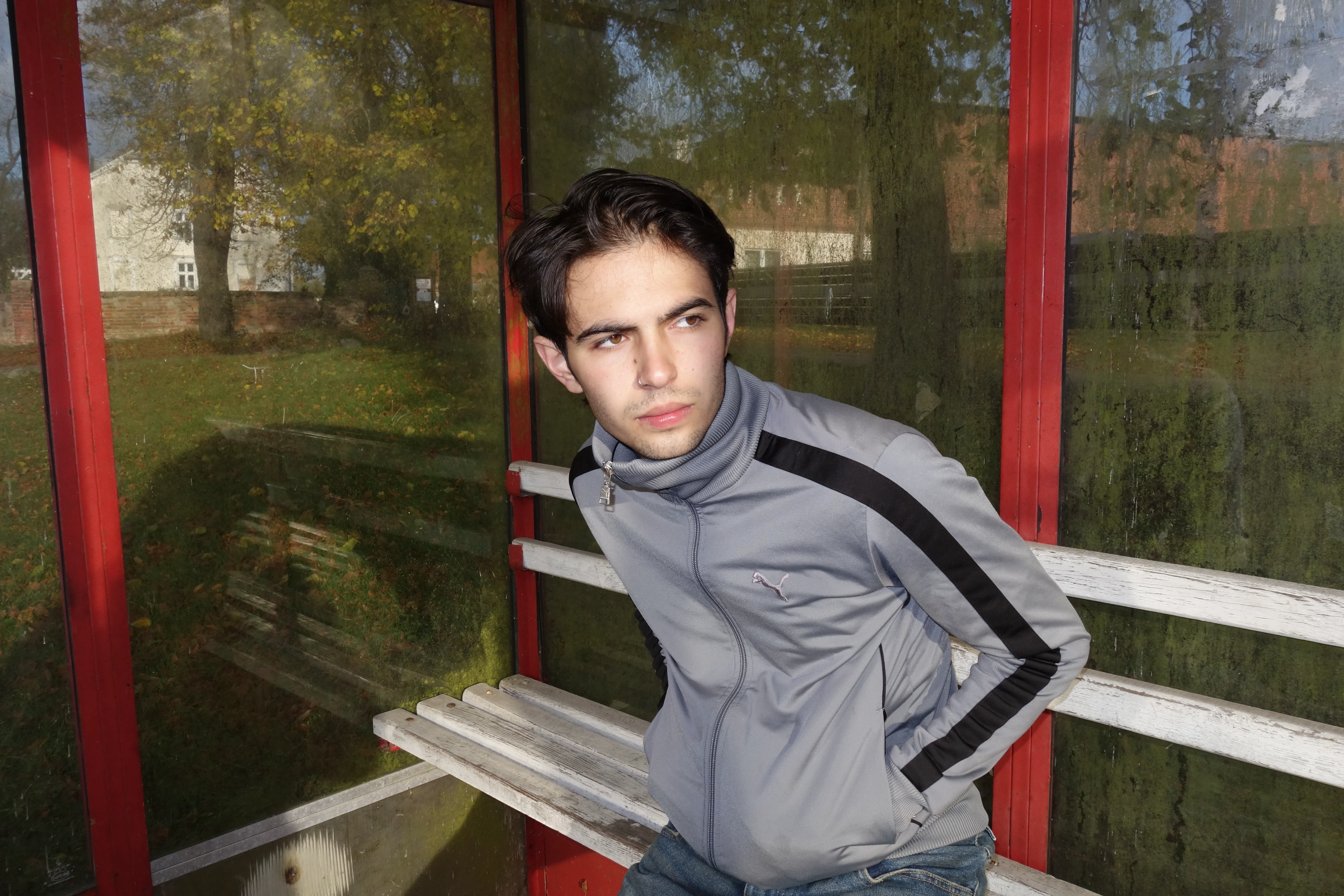 Ein junger Mann mit dunklen Haaren und grauer Trainingsjacke wartet in einer Buswartehalle.