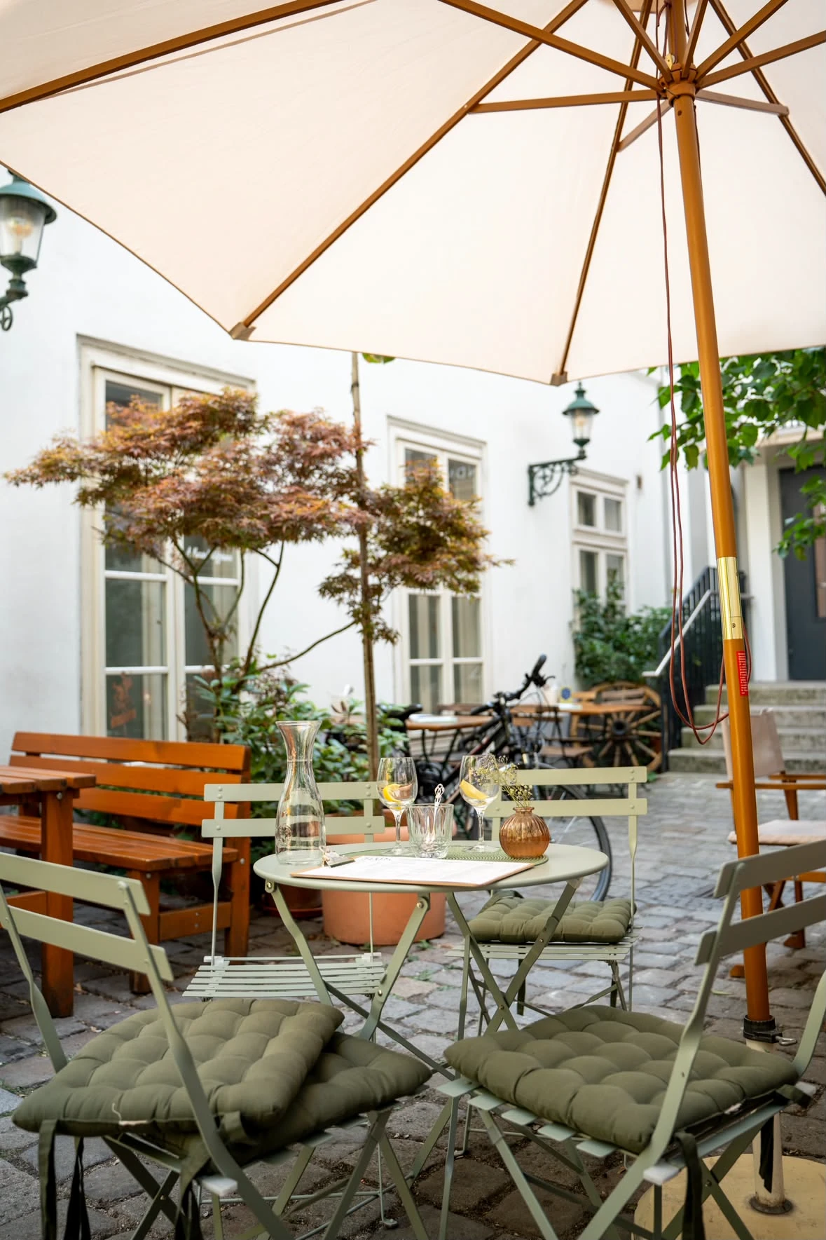 Gemütlicher Außenbereich eines Cafés mit einem gedeckten Tisch, grünen Stühlen und einem großen Sonnenschirm im Innenhof.