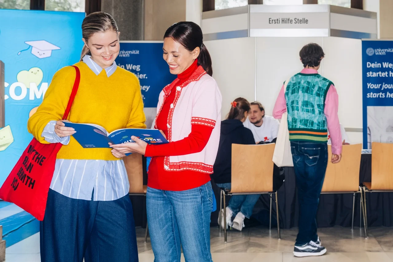 Zwei junge Studentinnen schauen sich auf dem Campus der Universität Wien eine Willkommensbroschüre an einer Infostand-Messe an.