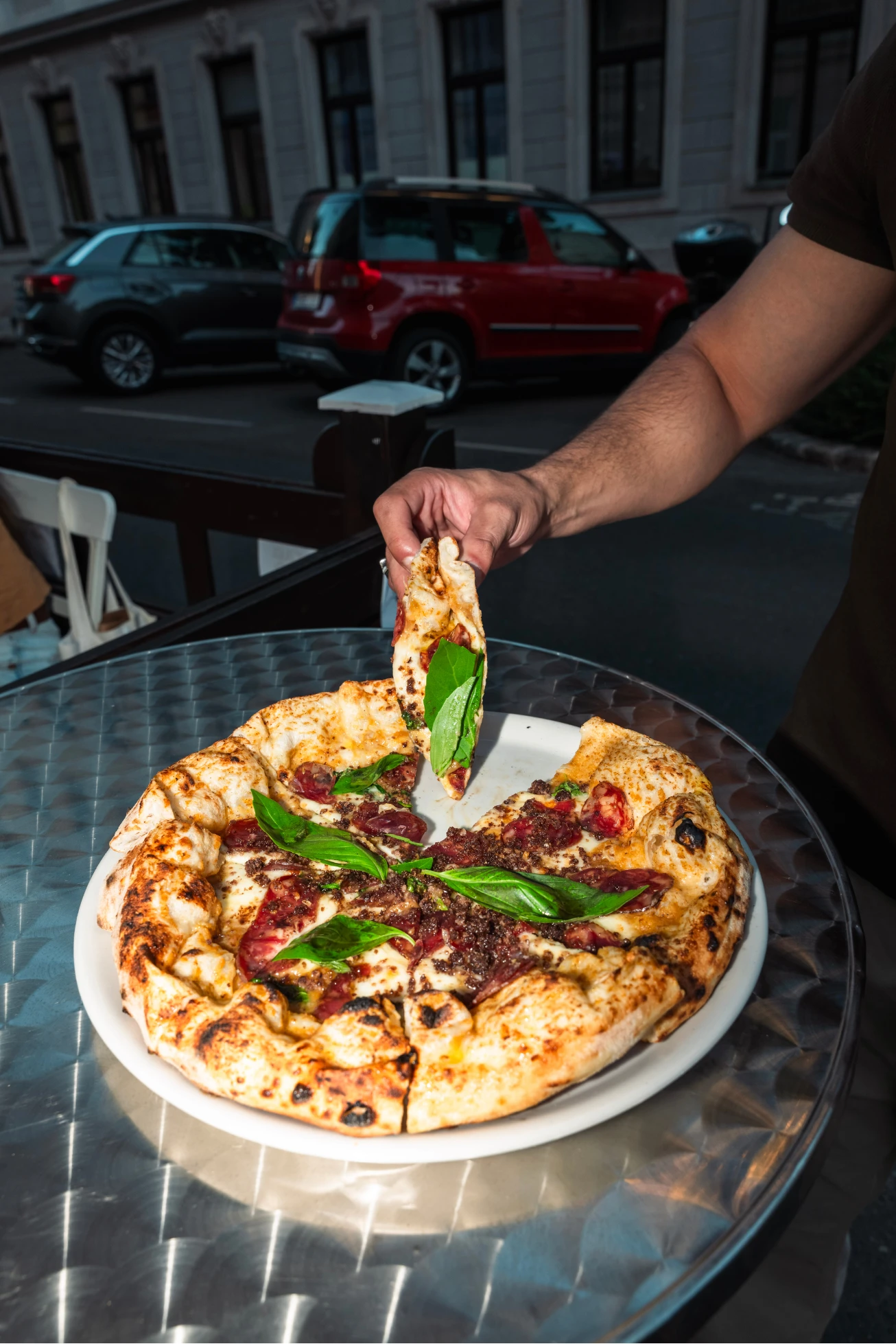 Eine Person hebt ein Stück Pizza mit Salami und Basilikum von einem Teller auf einem Tisch im Freien.
