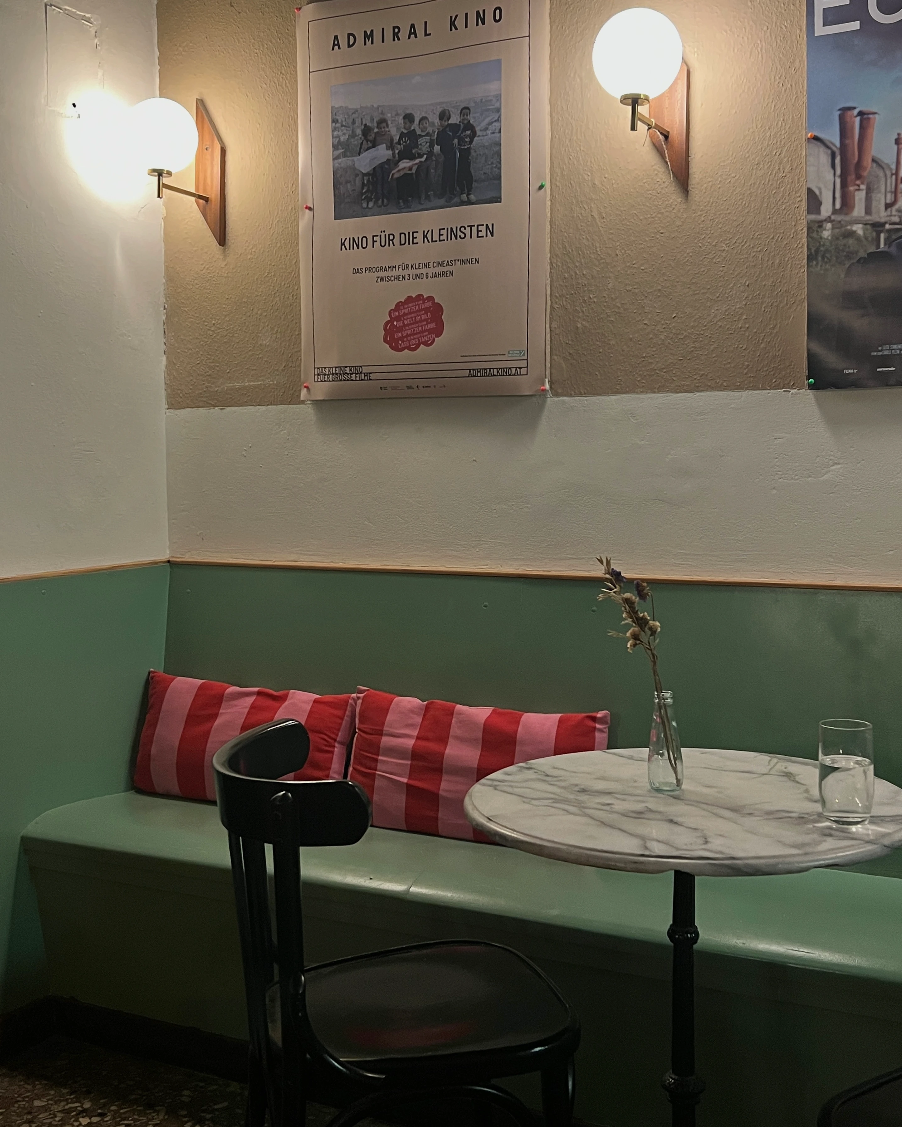 Gemütliche Sitzecke in einem Café mit einer grünen Bank, rotem Kissen, einem runden Marmortisch und Wandplakaten.