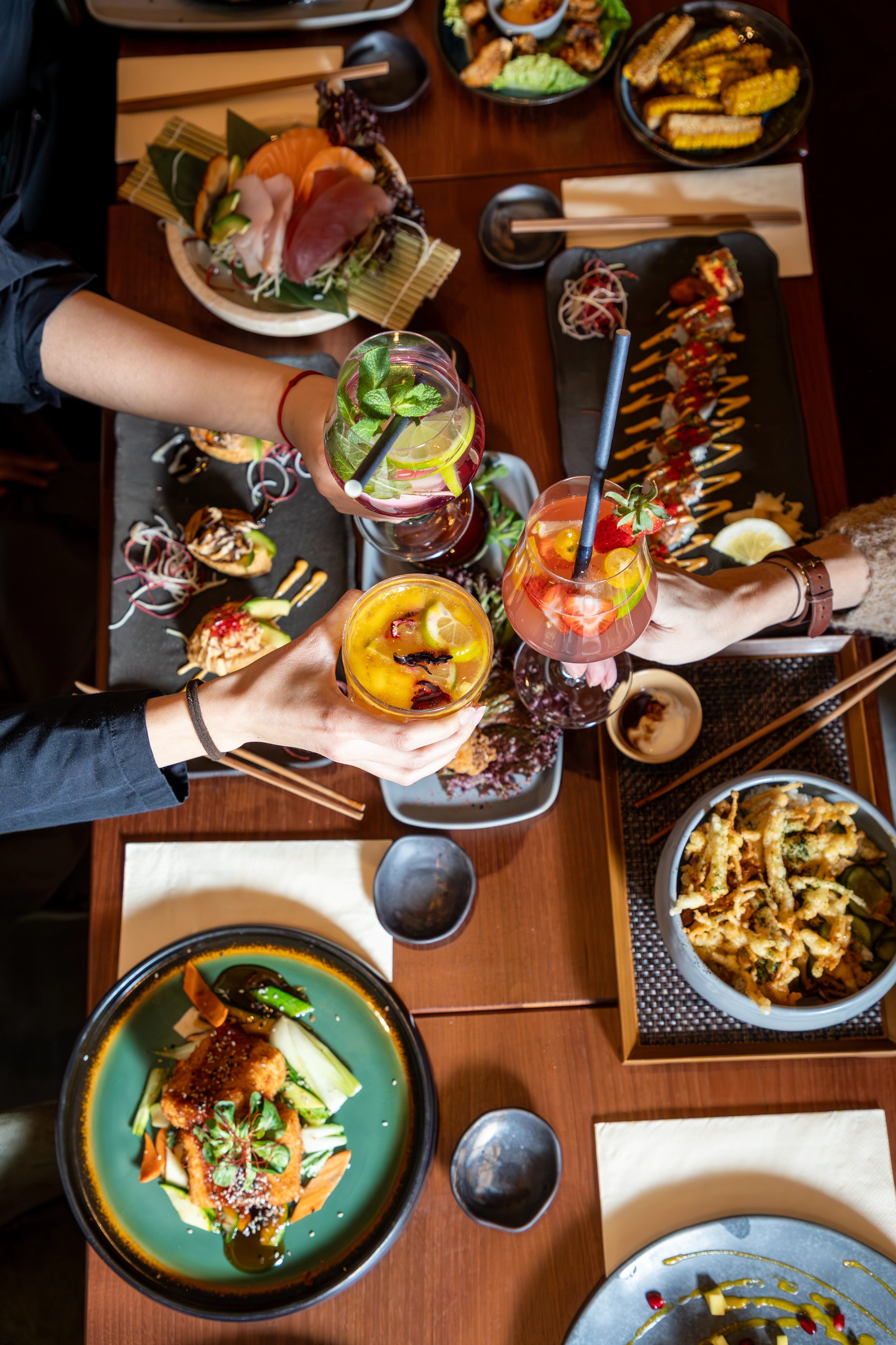 Draufsicht auf einen voll gedeckten Tisch mit verschiedenen asiatischen Gerichten und anstoßenden Cocktails.