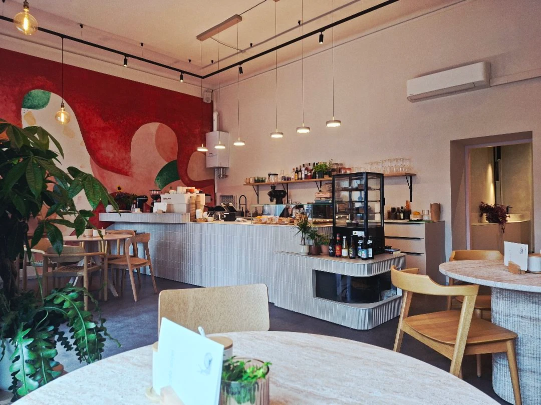 Modernes Café mit heller Theke, runden Tischen, Holzstühlen und einer großen roten Wandmalerei im Innenraum.