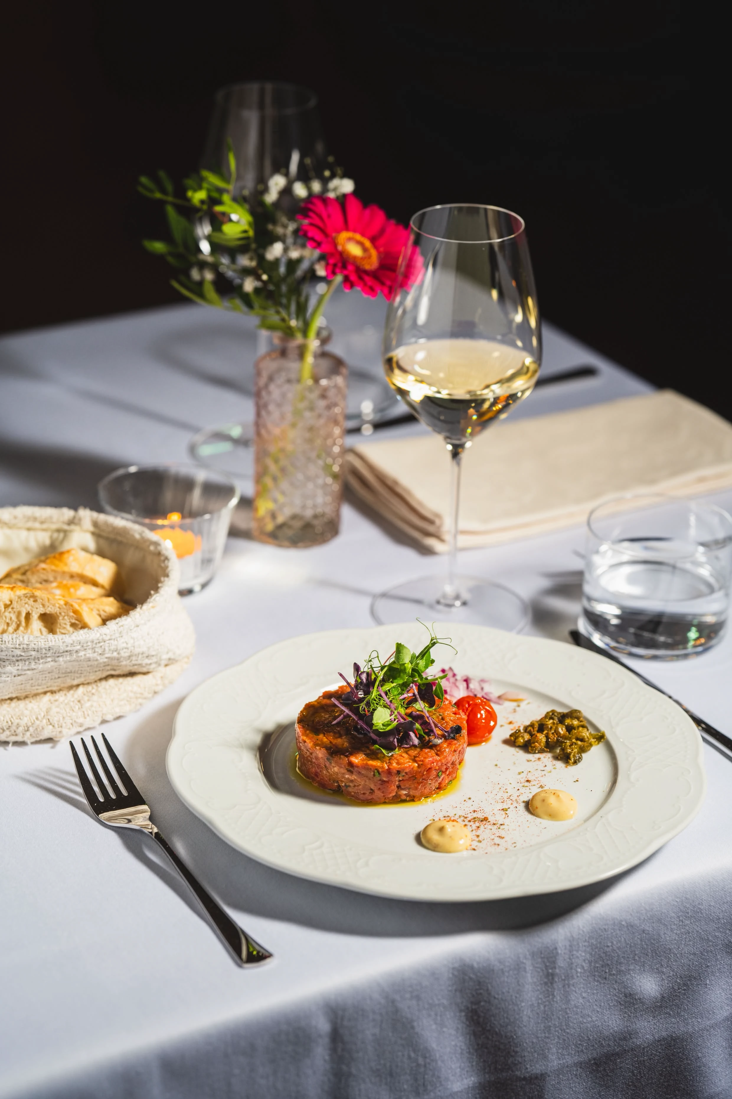 Elegantes Steak Tatar serviert auf einem weißen Teller mit Beilagen, begleitet von einem Glas Weißwein und einer Blumendekoration auf einem gedeckten Tisch im Restaurant.