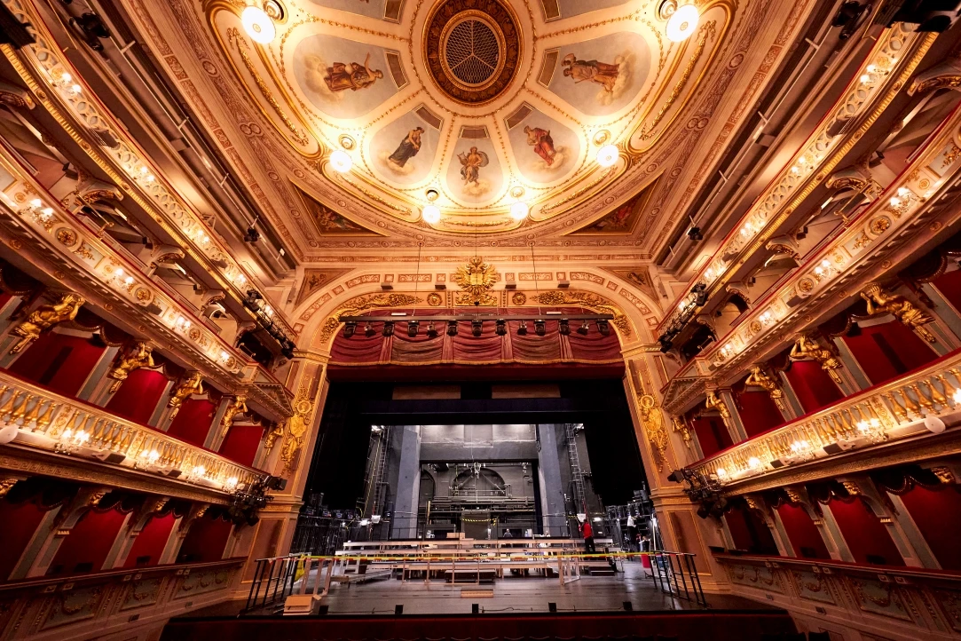 Prunkvoller Zuschauerraum eines historischen Theaters mit kunstvollem Deckengemälde, goldenen Verzierungen, roten Logen und Blick auf die leere Bühne.