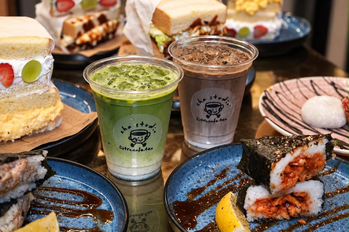 Tisch mit verschiedenen Getränken wie Matcha und Schokolade sowie japanischen Sandwiches und Onigiri in einem Café.