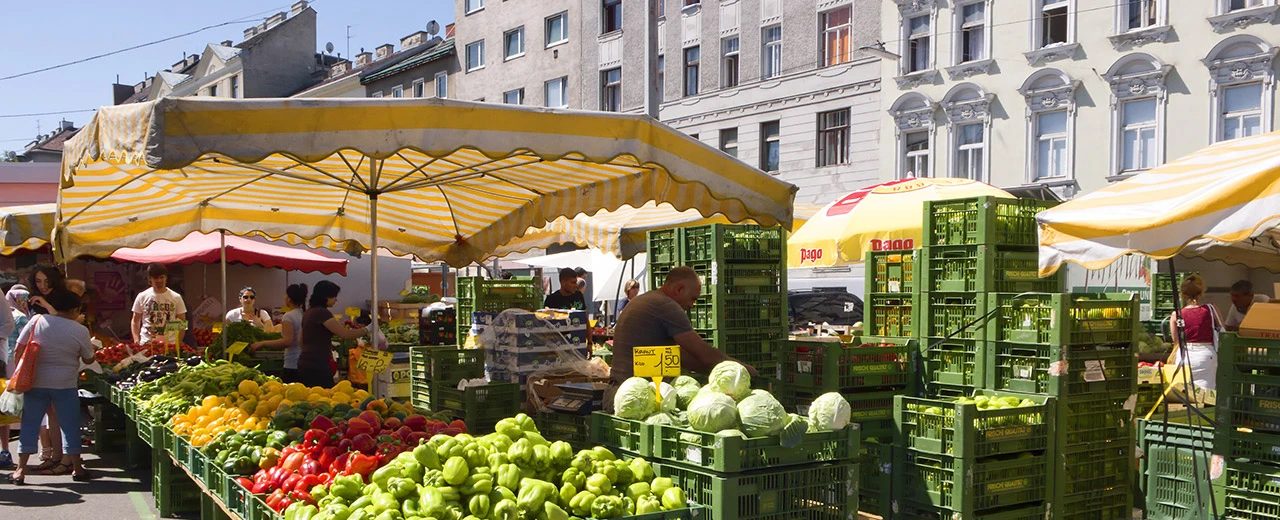 Lebhafter Marktstand in Wien mit buntem Obst und Gemüse unter gelben und weißen Sonnenschirmen.