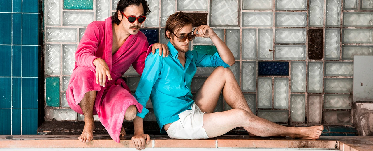 © Julian & der Fux Zwei Männer, einer in rosa Bademantel, der andere in türkisfarbenem Hemd und weißen Shorts, sitzen am Poolrand vor einer Glasbausteinfliese. Mit Adobe Photoshop CS6 erstellt.