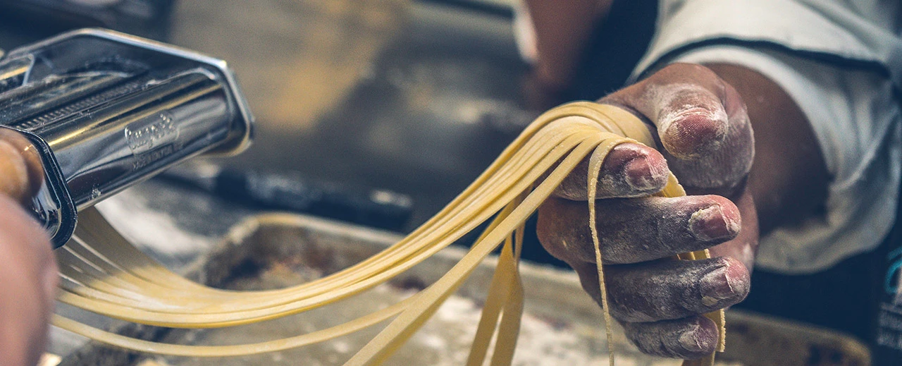 Hände eines Kochs halten frisch gemachte Pasta-Bänder nach dem Passieren durch eine Nudelmaschine.
