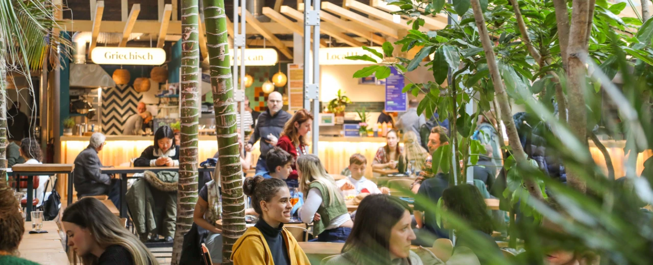 © Gleis//Garten Lebhafte Szene in einem grünen Food Court mit vielen Menschen an Tischen. Verschiedene Essensstände und viel Pflanzen. Gleisgarten.