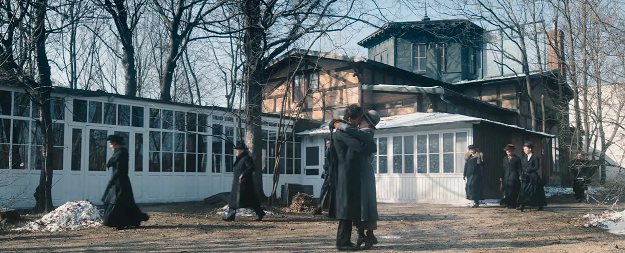 Liebespaar umarmt sich vor einem historischen Gebäude; mehrere Personen in dunkler Kleidung im Hintergrund, Winterlandschaft.