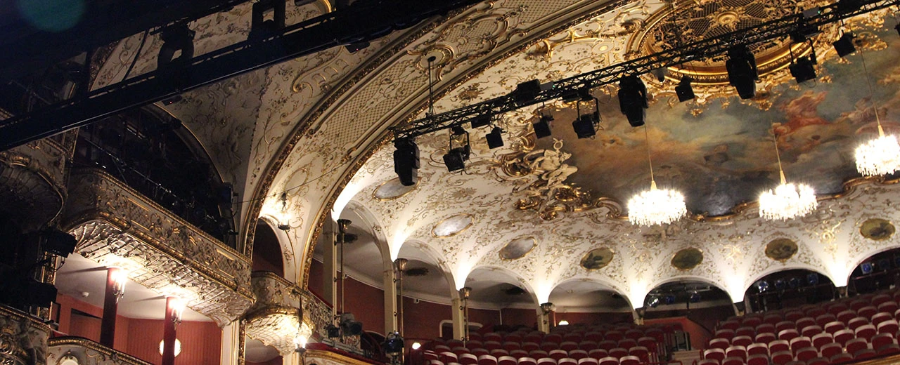 Prachtvoller Theatersaal mit Stuckdecke, Kronleuchtern & roten Sitzen. Historische Architektur & moderne Bühnenbeleuchtung.