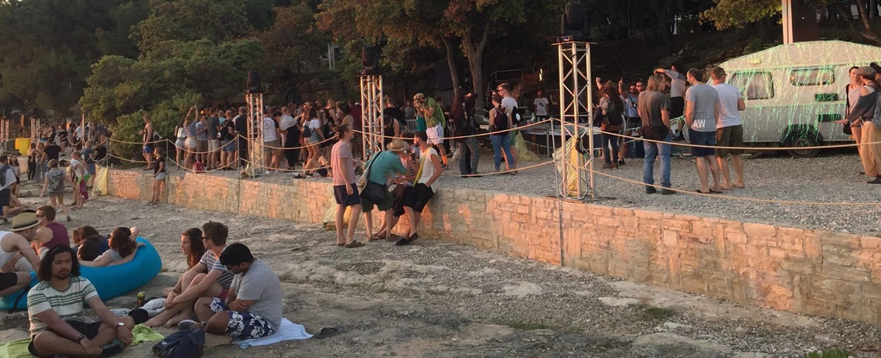 Open-Air-Konzert an einem Ufer mit vielen Zuschauern, einer Steinmauer und einem Wohnmobil im Hintergrund. Sommerliche Stimmung.