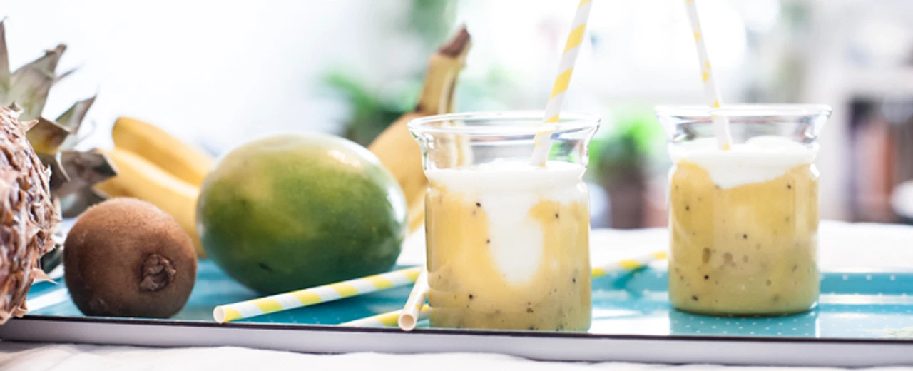 Zwei Gläser fruchtiger Smoothie mit Mango, Ananas, Banane und Kiwi auf einer türkisfarbenen Platte. Mit Lightroom bearbeitet.