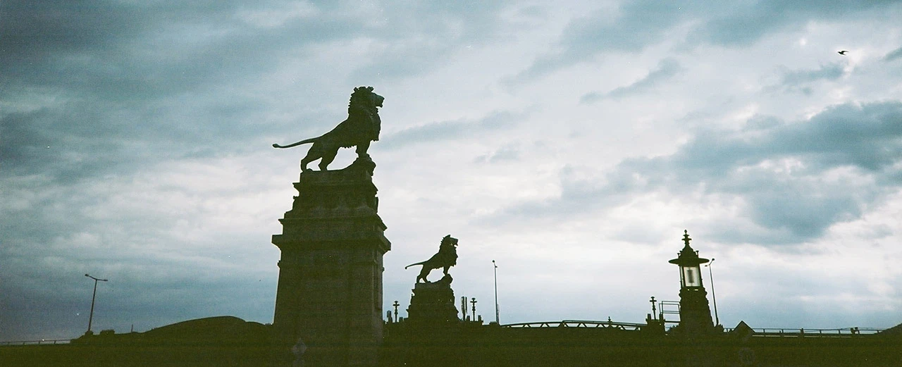 Zwei Löwenstatuen auf hohen Sockeln vor einem bewölkten Himmel.  Hintergrund: Brücke und Laternen.