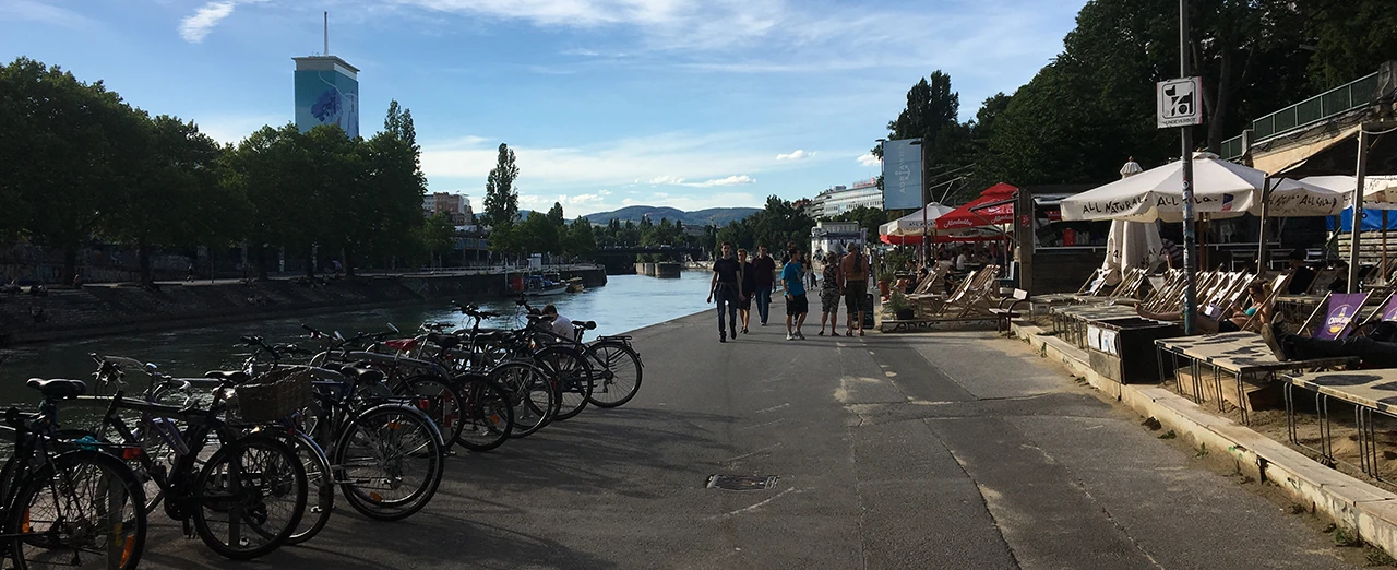 Fahrradständer, Fluss, Spaziergänger, Restaurants & Bars mit Sonnenschirmen an einem sonnigen Tag.