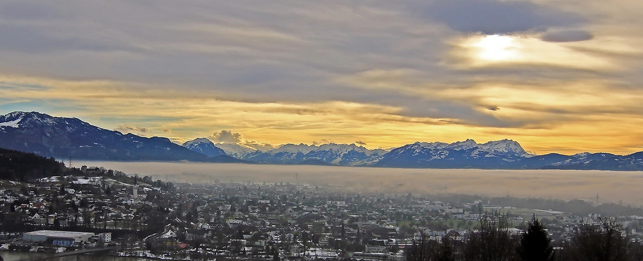 © Dornbirn by Smaragd auf Pixabay Panorama einer Stadt im Alpenvorland bei Sonnenuntergang, umgeben von Nebel und schneebedeckten Bergen.  Erstellt mit Adobe Photoshop.
