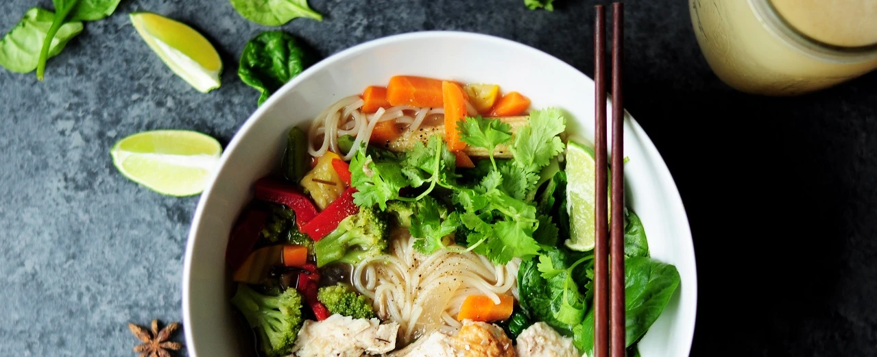 Schüssel asiatische Nudelsuppe mit Huhn, Gemüse & Koriander. Limetten & Essstäbchen.