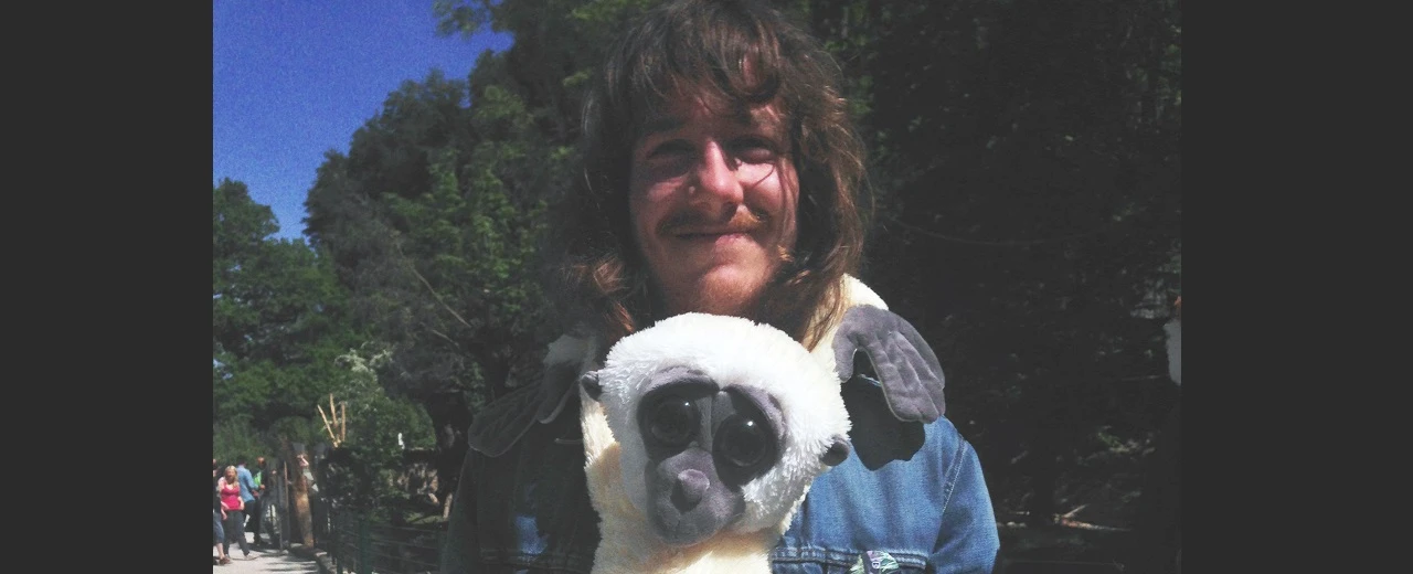 Junger Mann mit langem Haar lächelt und trägt ein Lemur-Plüschtier.  Erstellt mit Adobe Photoshop CS3.