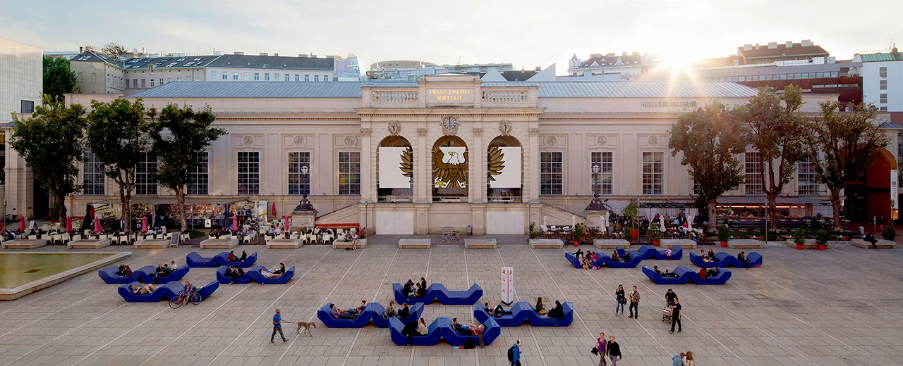 Franz-Josefs-Bahnhof in Wien: Menschen entspannen auf modernen blauen Bänken vor dem historischen Gebäude bei Sonnenuntergang.