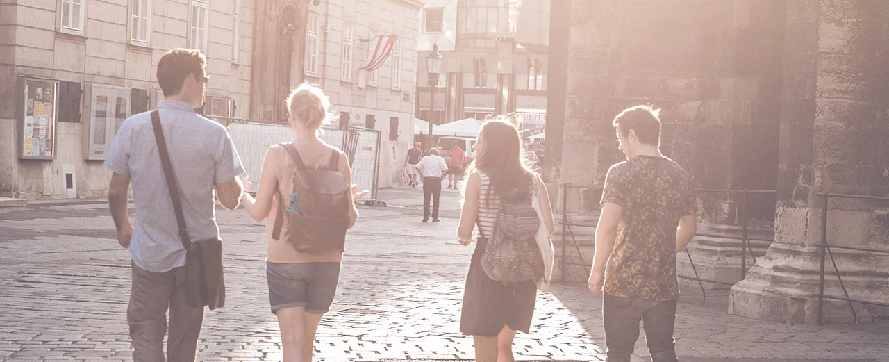 Vier junge Erwachsene spazieren auf einem gepflasterten Platz in einer historischen europäischen Stadt. Warme, helle Farben, sonniger Tag.