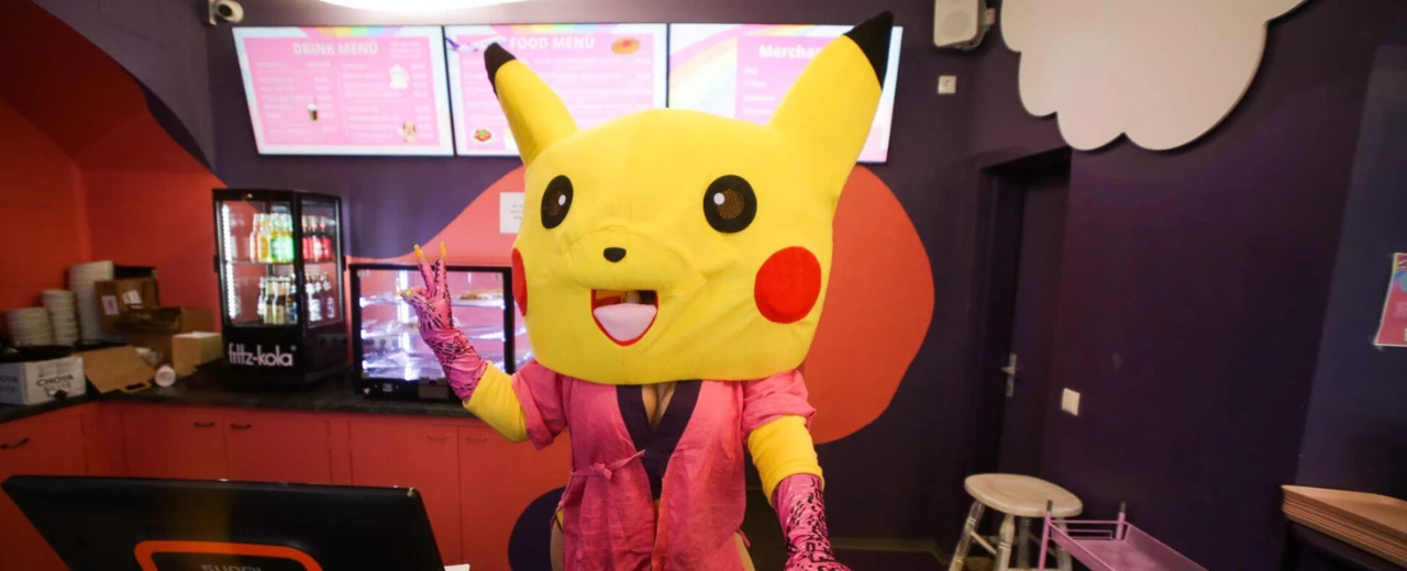 Pikachu-Maskottchen in einem Café, macht Peace-Zeichen. Speisekarten und Theke im Hintergrund.