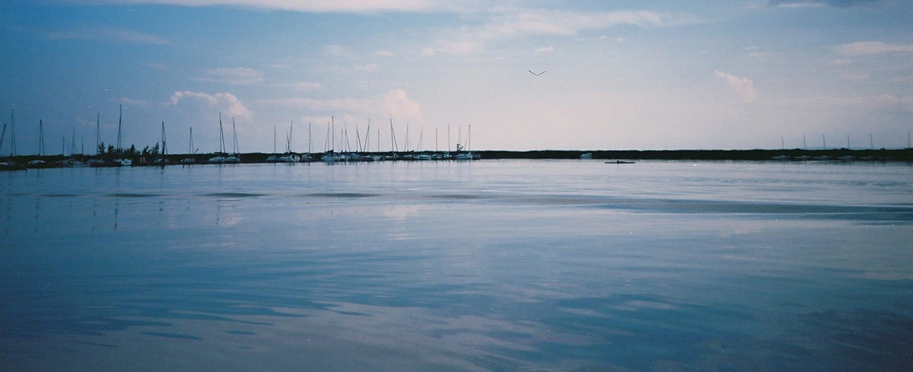 © Miriam Kummer Ruhige Wasseroberfläche, Segelboote im Hintergrund, leicht bewölkter Himmel.