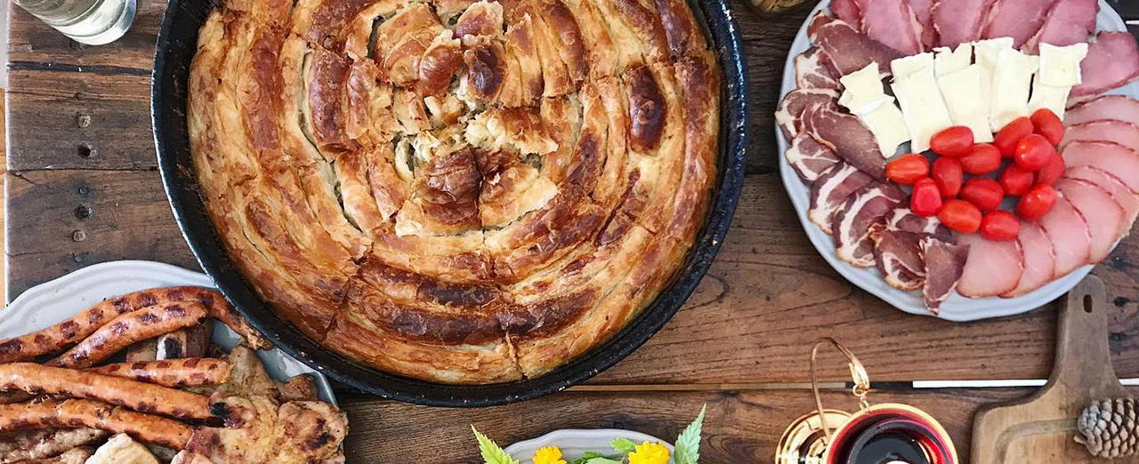 Bild mit verschiedenen Speisen: Burek, Aufschnitt, Käse, Tomaten, gegrilltes Fleisch und Würstchen auf einem Holztisch. Mit Windows Foto-Editor bearbeitet.