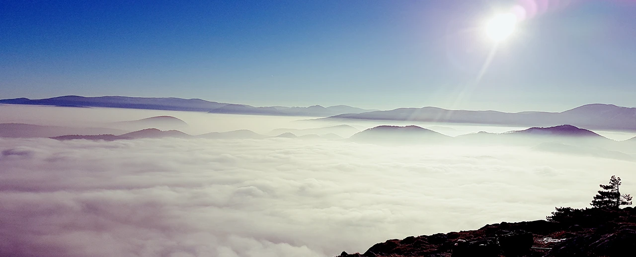 ©  Atemberaubende Aussicht von einem Berg auf ein Meer aus Wolken, Berge im Hintergrund, strahlende Sonne.